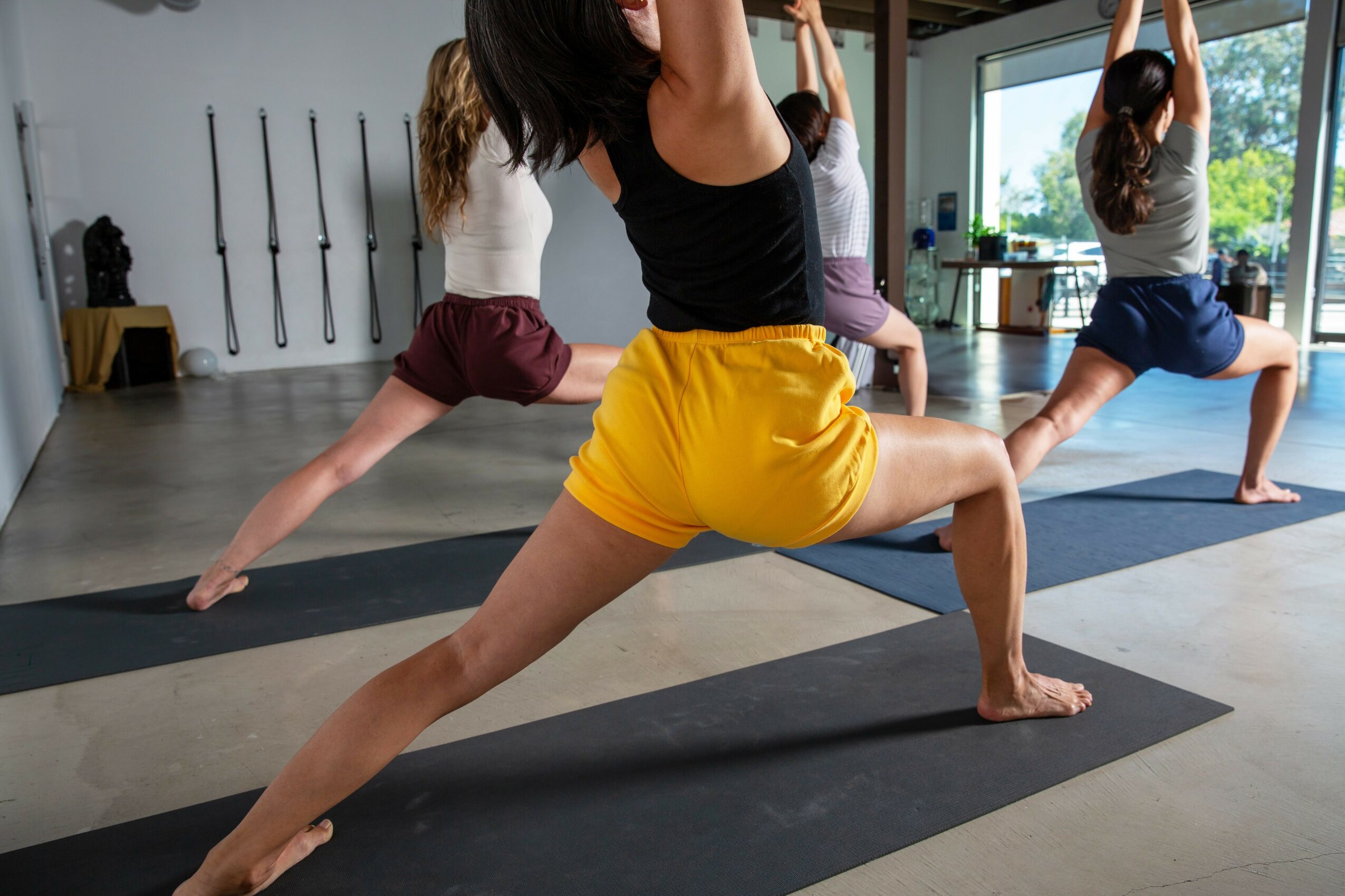 Group of people practicing yoga in a studio, performing Warrior pose on mats, with large windows and sunlight.