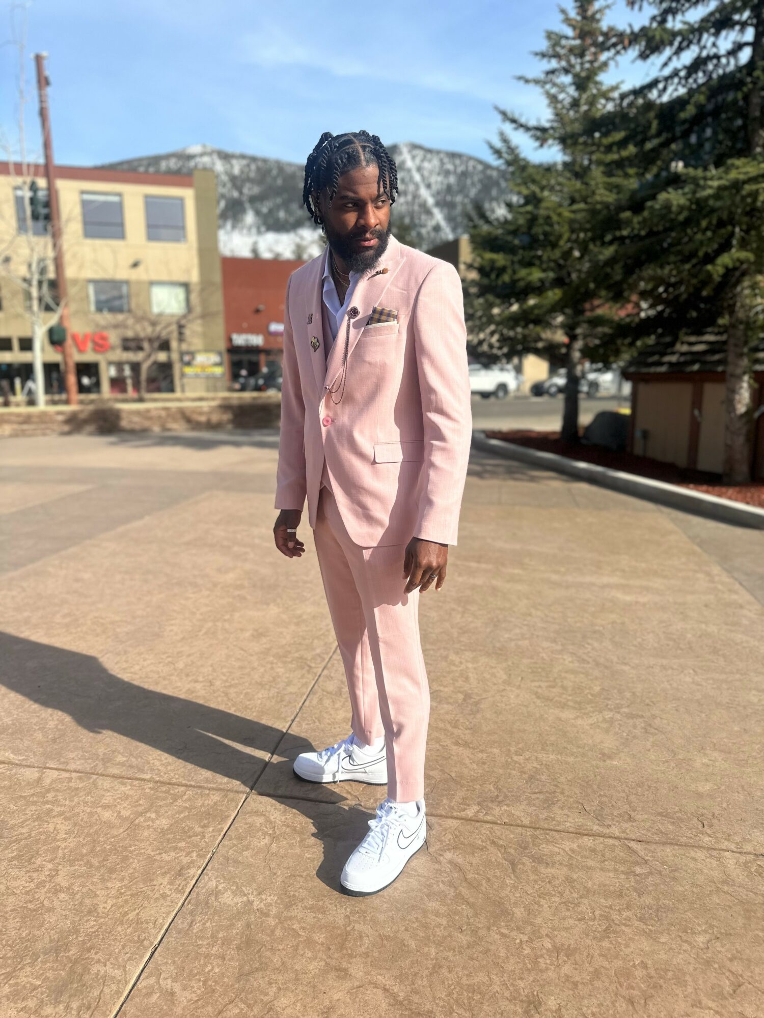 Person in a light pink suit standing outdoors on a paved area with buildings and trees in the background.