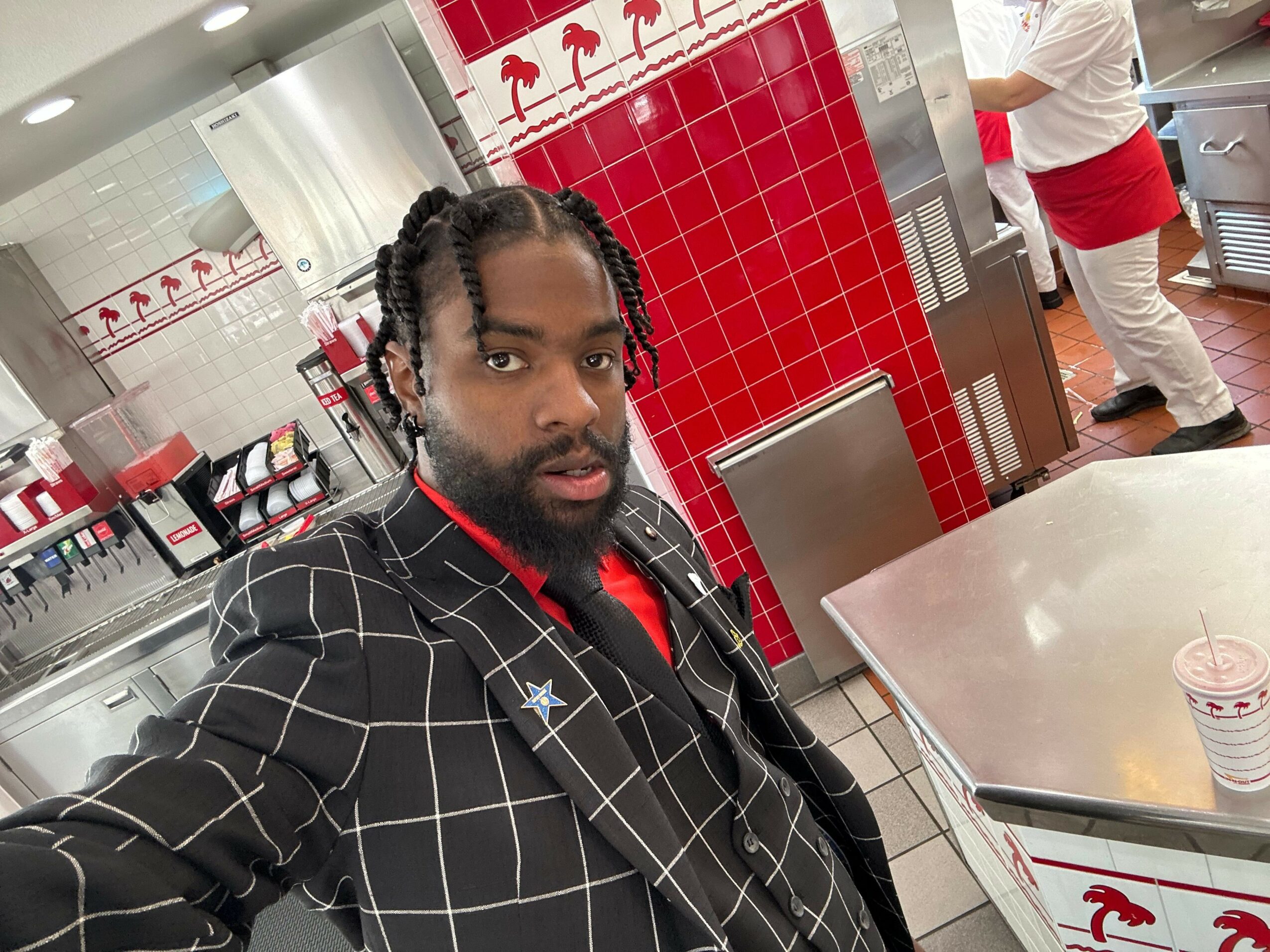 Person with braided hair and beard wearing a black jacket with white grid pattern, standing in a store with red and white tiles.