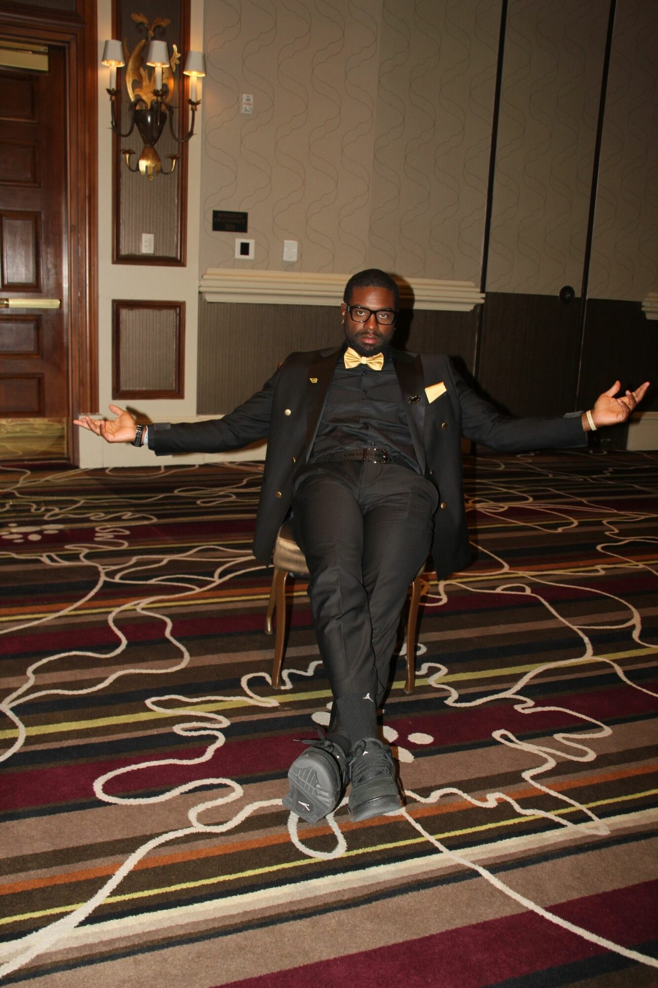 Person in a suit standing on a patterned carpet, with cords on the floor, in a room with wooden doors and wall decor.