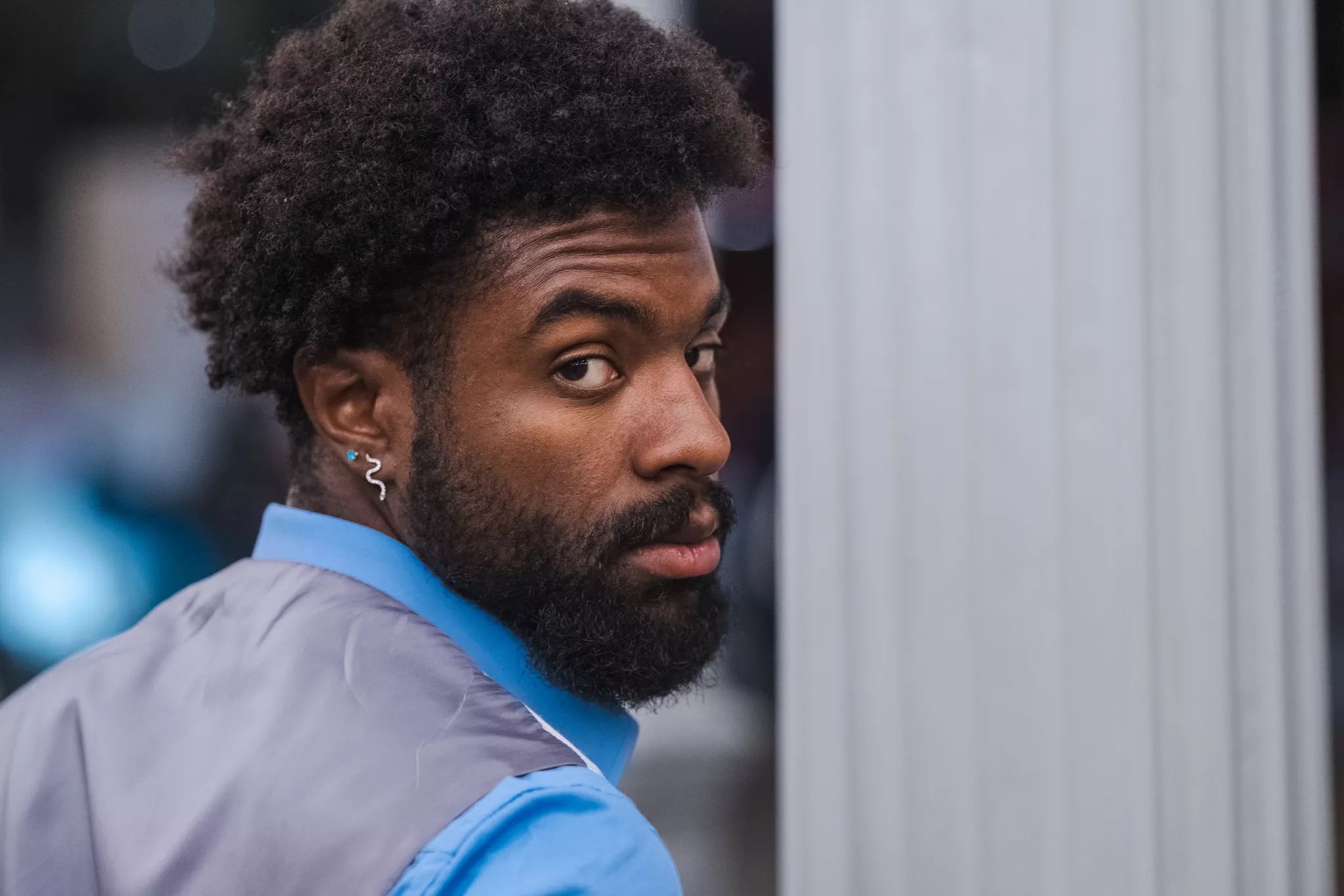 Portrait of a man with curly hair and beard, wearing a blue shirt and gray jacket, looking over his shoulder.