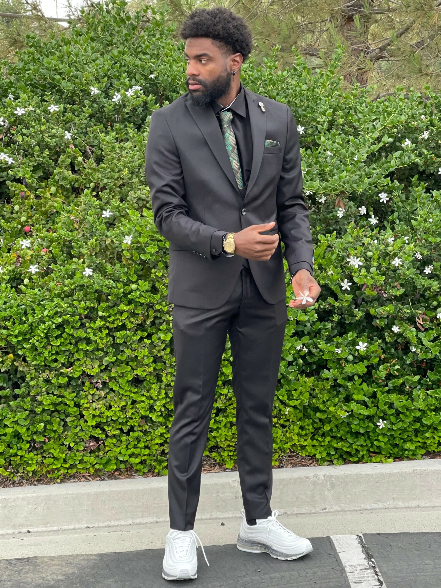 Man in a black suit with a green tie and white sneakers standing outdoors in front of green bushes.