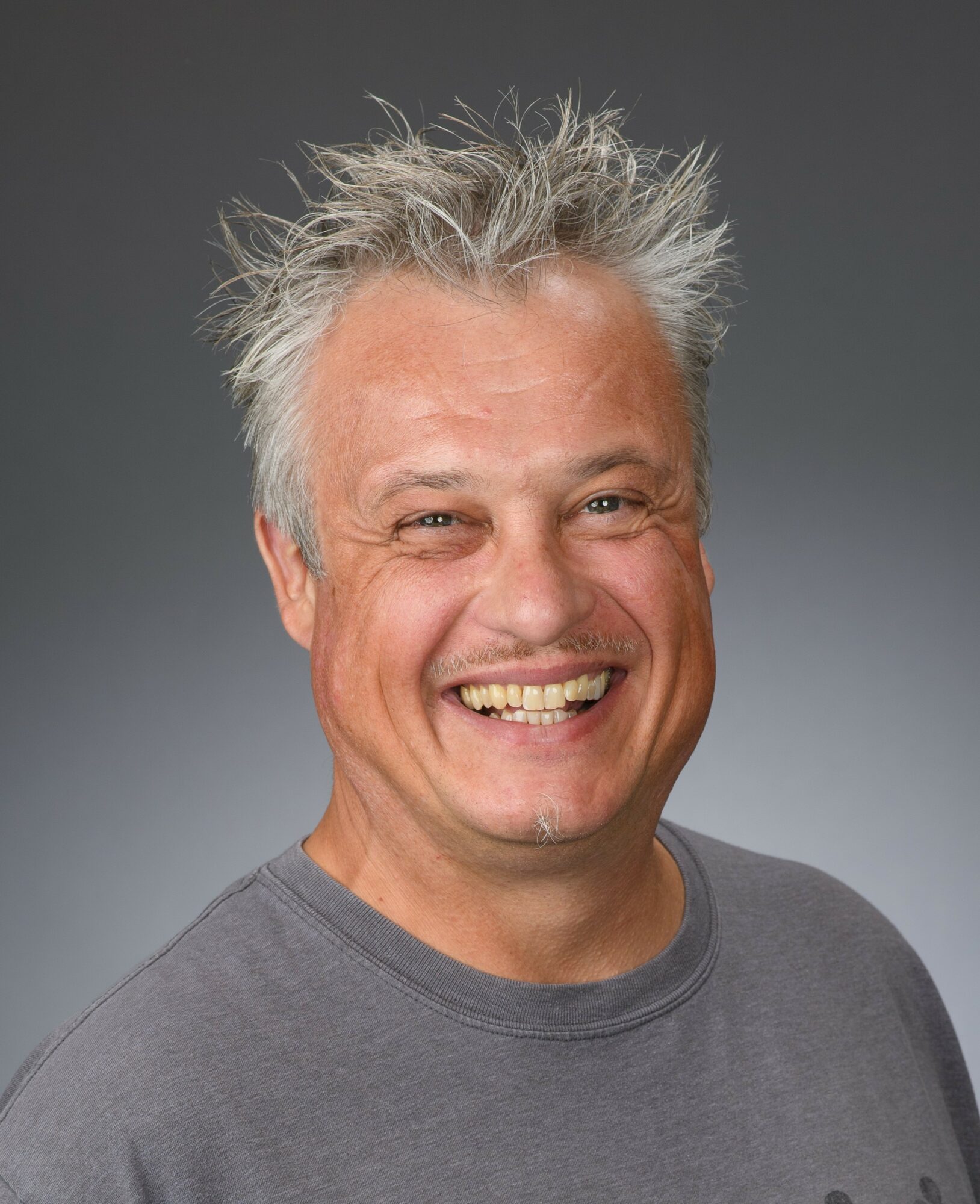 Smiling man with spiky gray hair wearing a gray shirt, facing forward, against a gray background.
