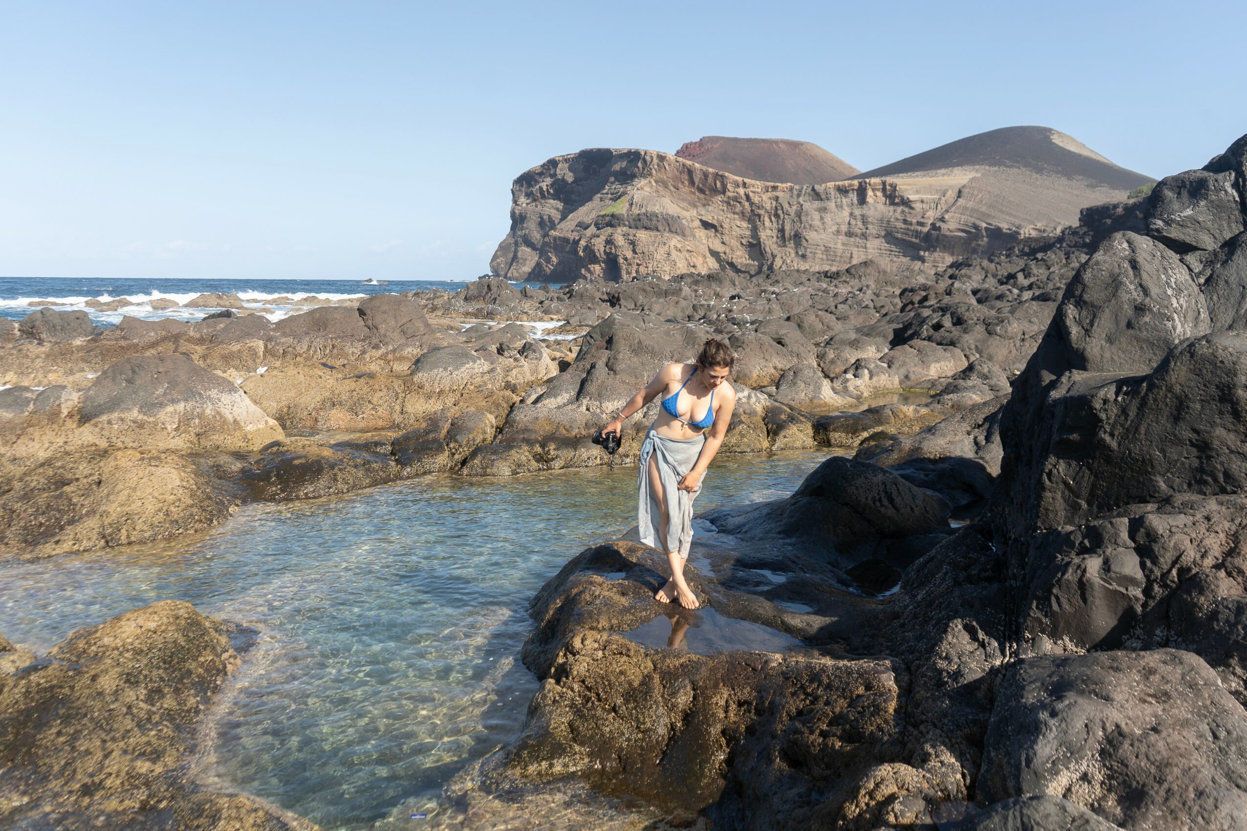 Person standing on rocks near water with cliffs and ocean in background, clear sky, daytime.