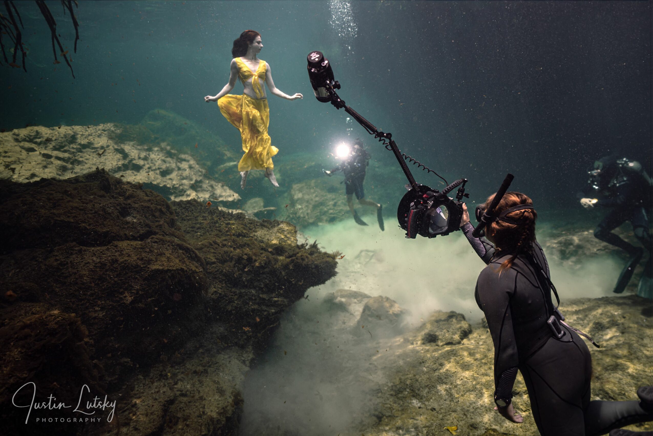 A woman in a yellow dress underwater holding a camera with a large light, surrounded by rocks and other divers.