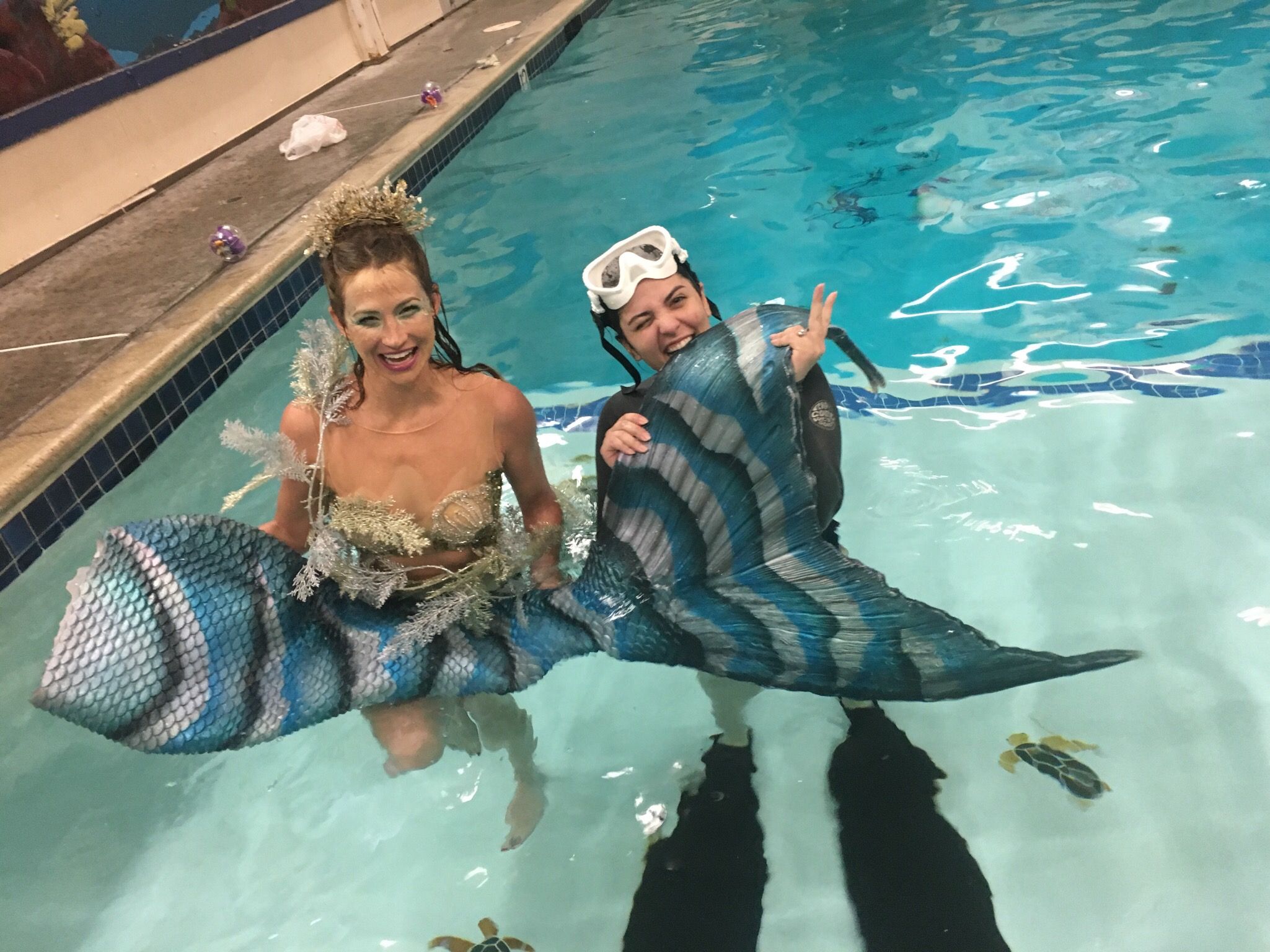 Two women in a swimming pool holding a large fish, one wearing goggles, smiling, and giving a thumbs-up, with water and pool edge visible.