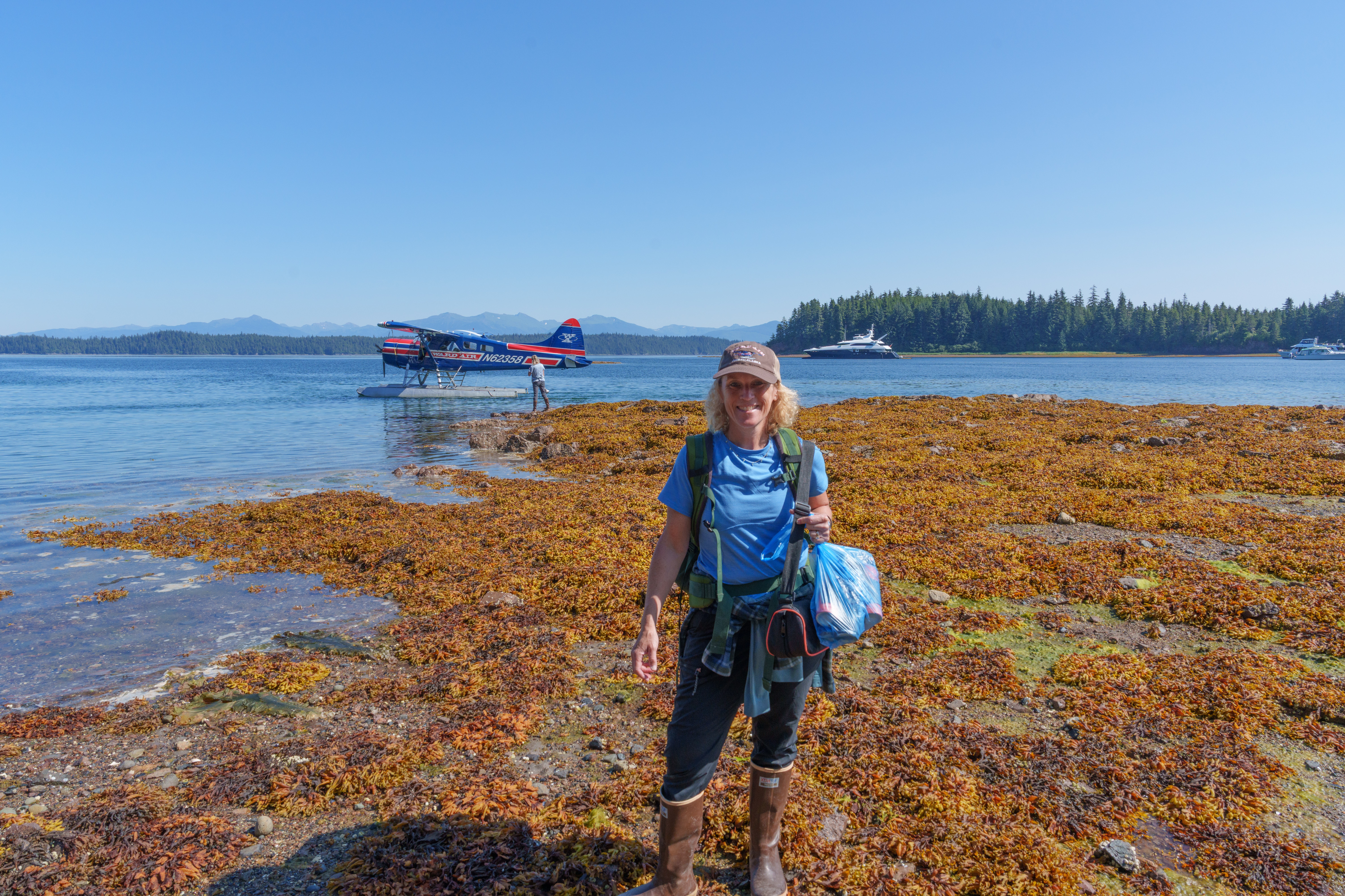Person standing on a rocky shoreline with a backpack, holding a bag, near water with a boat and trees in background.