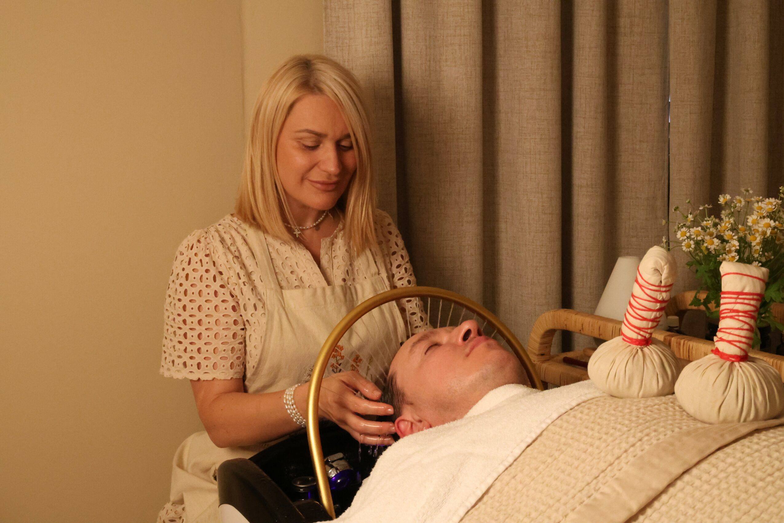 Woman with blonde hair smiling while tending to a person lying in bed, with curtains and decorative items in the background.
