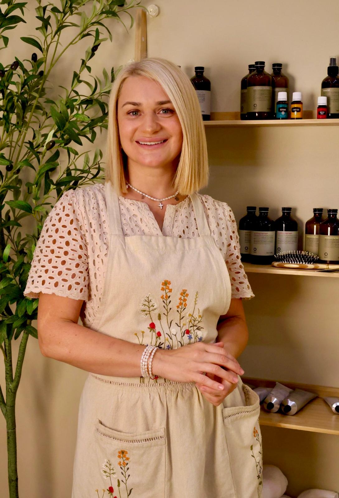 Smiling woman with blonde hair in a cream apron, standing in a room with shelves of bottles and plants.
