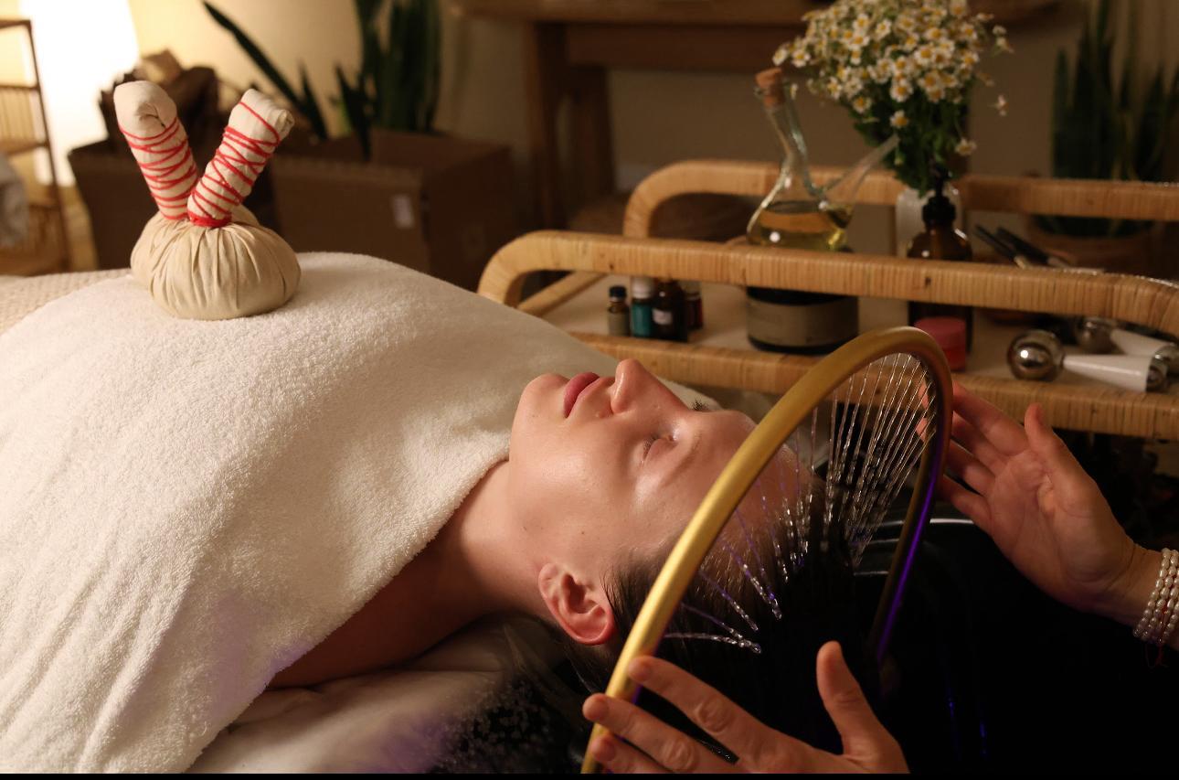 Person lying on a massage table with a towel covering their face, holding a colorful fan, in a cozy room with plants and flowers.