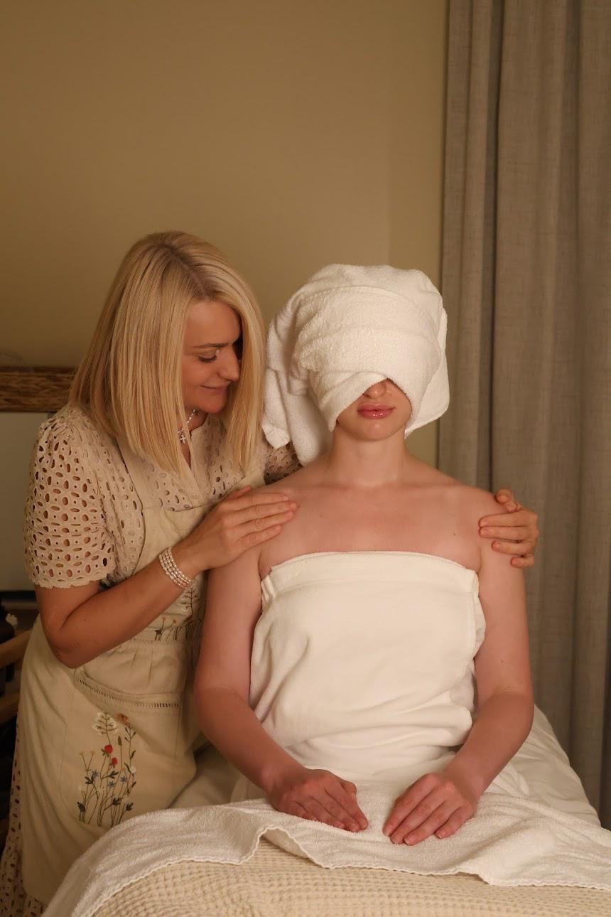 Person with a towel over their eyes sitting on a bed, another person standing beside them adjusting their towel.