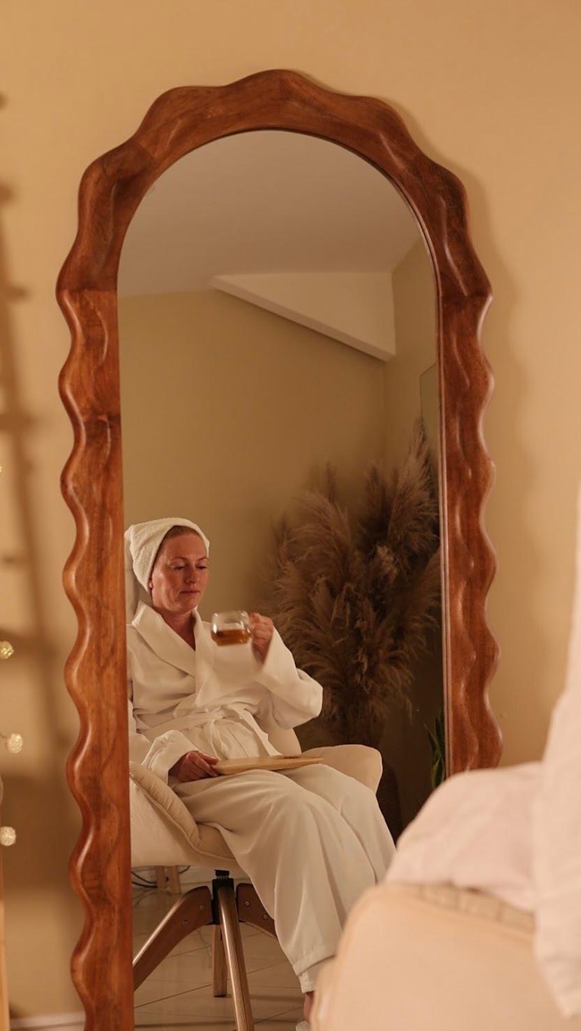 Person sitting in front of a tall, decorative mirror taking a selfie, wearing light-colored clothing, with a plant in the background.