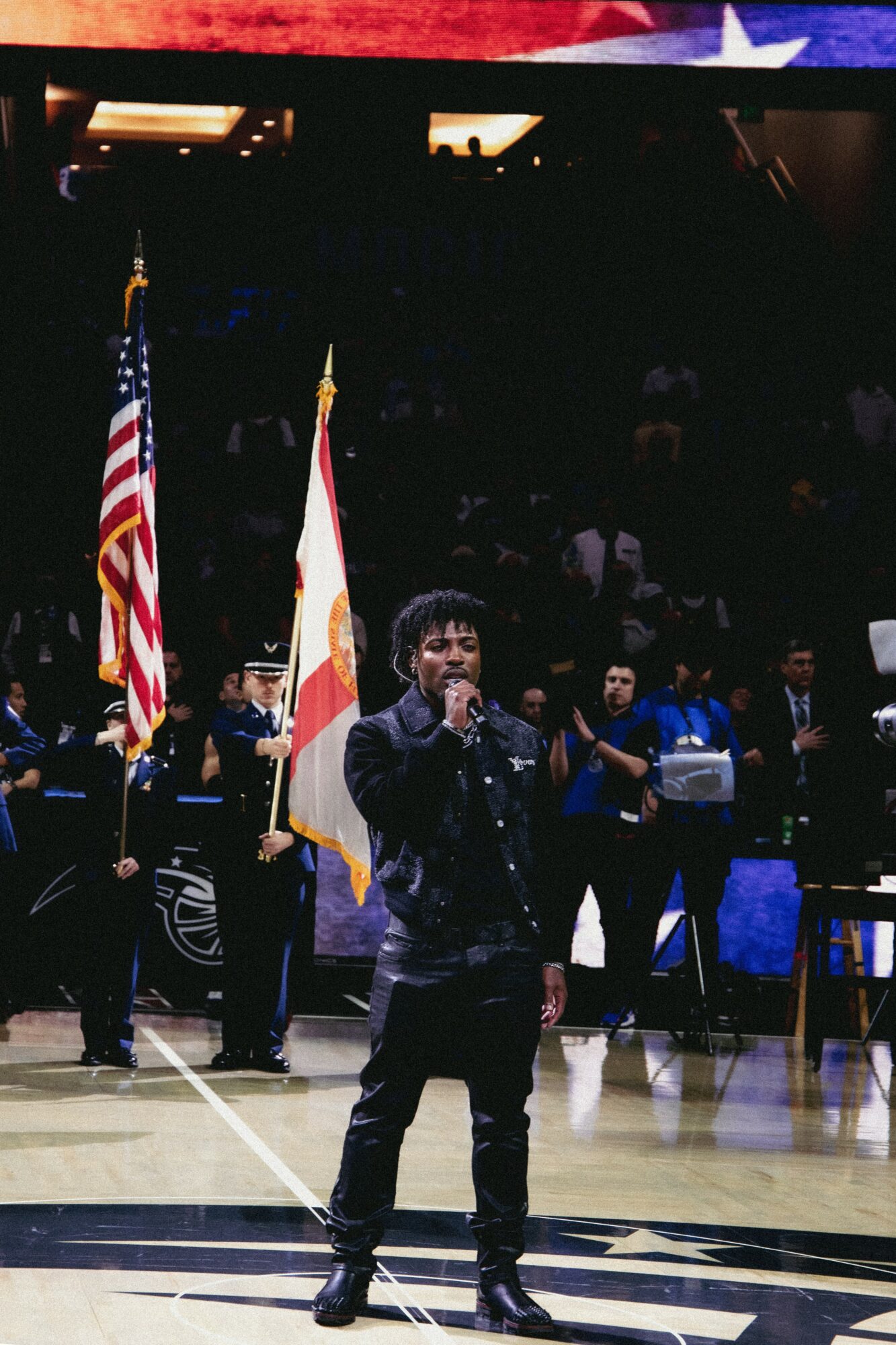Person singing on stage with American and Florida flags behind, audience in background, indoor sports arena setting.