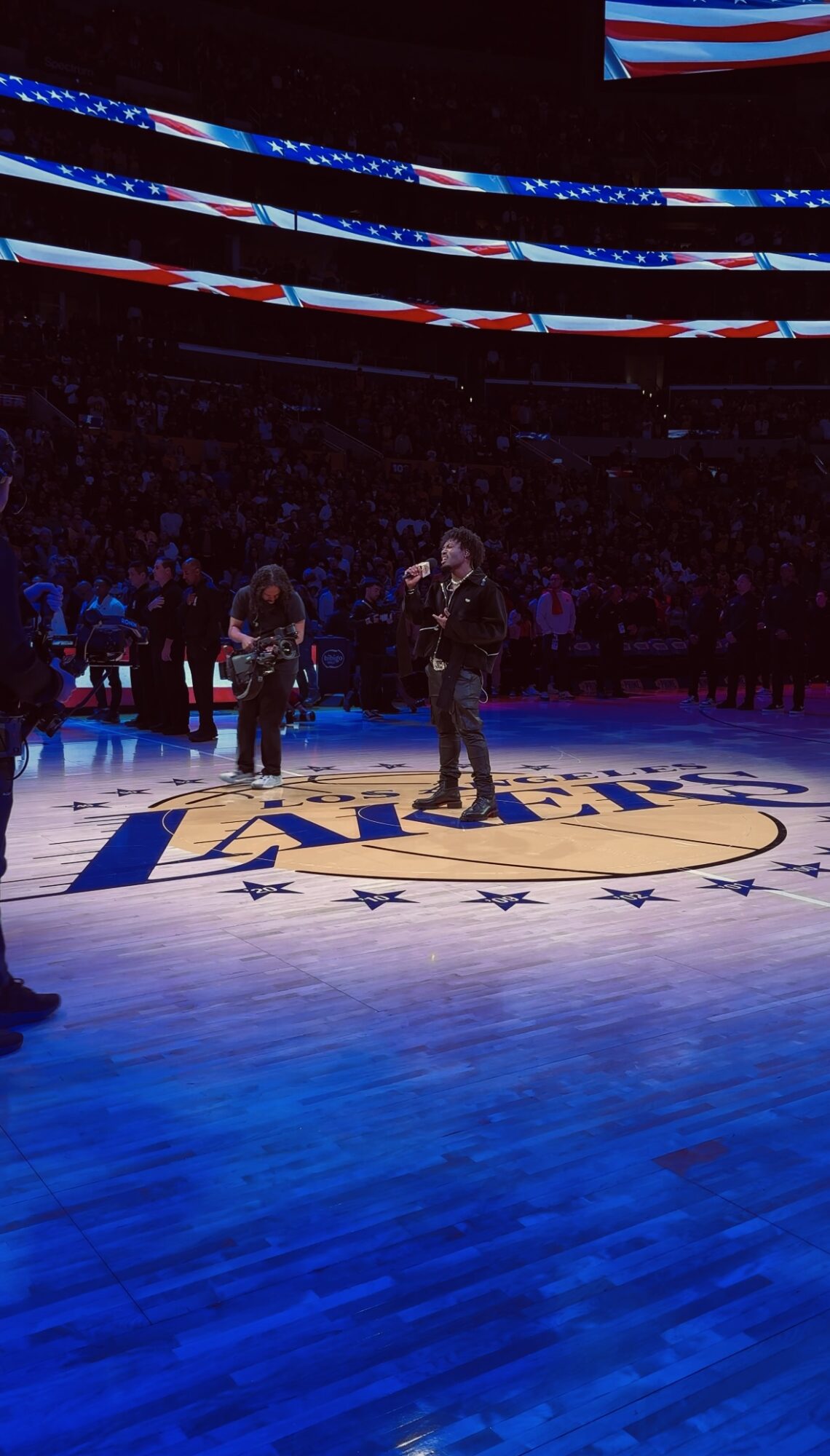 People standing on a basketball court with a large logo, surrounded by banners and an audience, in a sports arena.