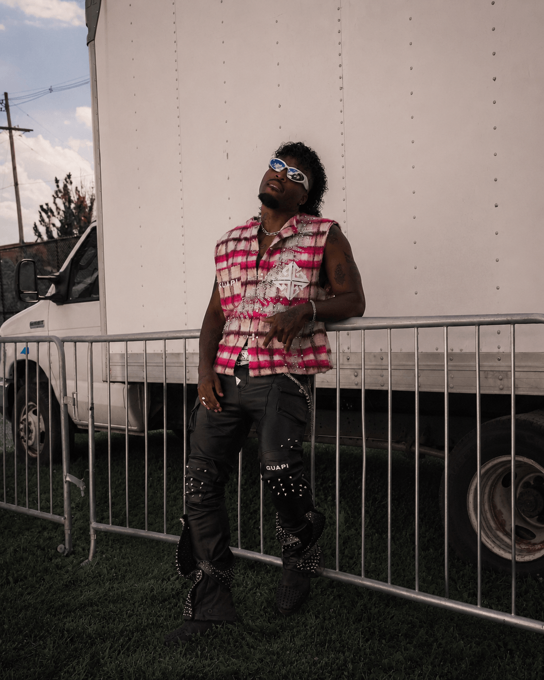 Person with sunglasses and a sleeveless shirt leaning against a metal fence outdoors, with a large white truck behind them.