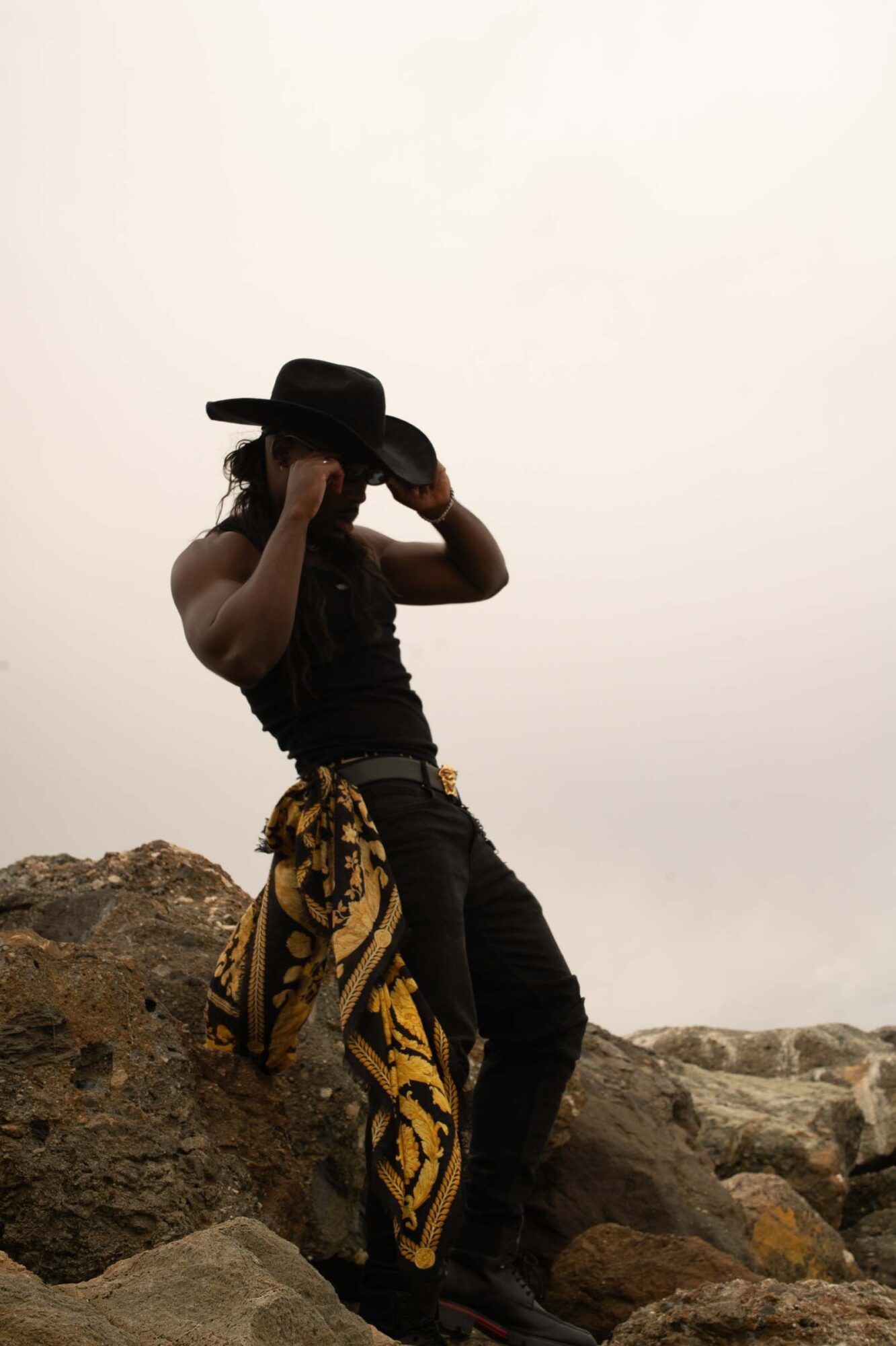 Person standing on rocks, wearing a hat and black clothing, holding the hat with both hands, with a plain sky background.
