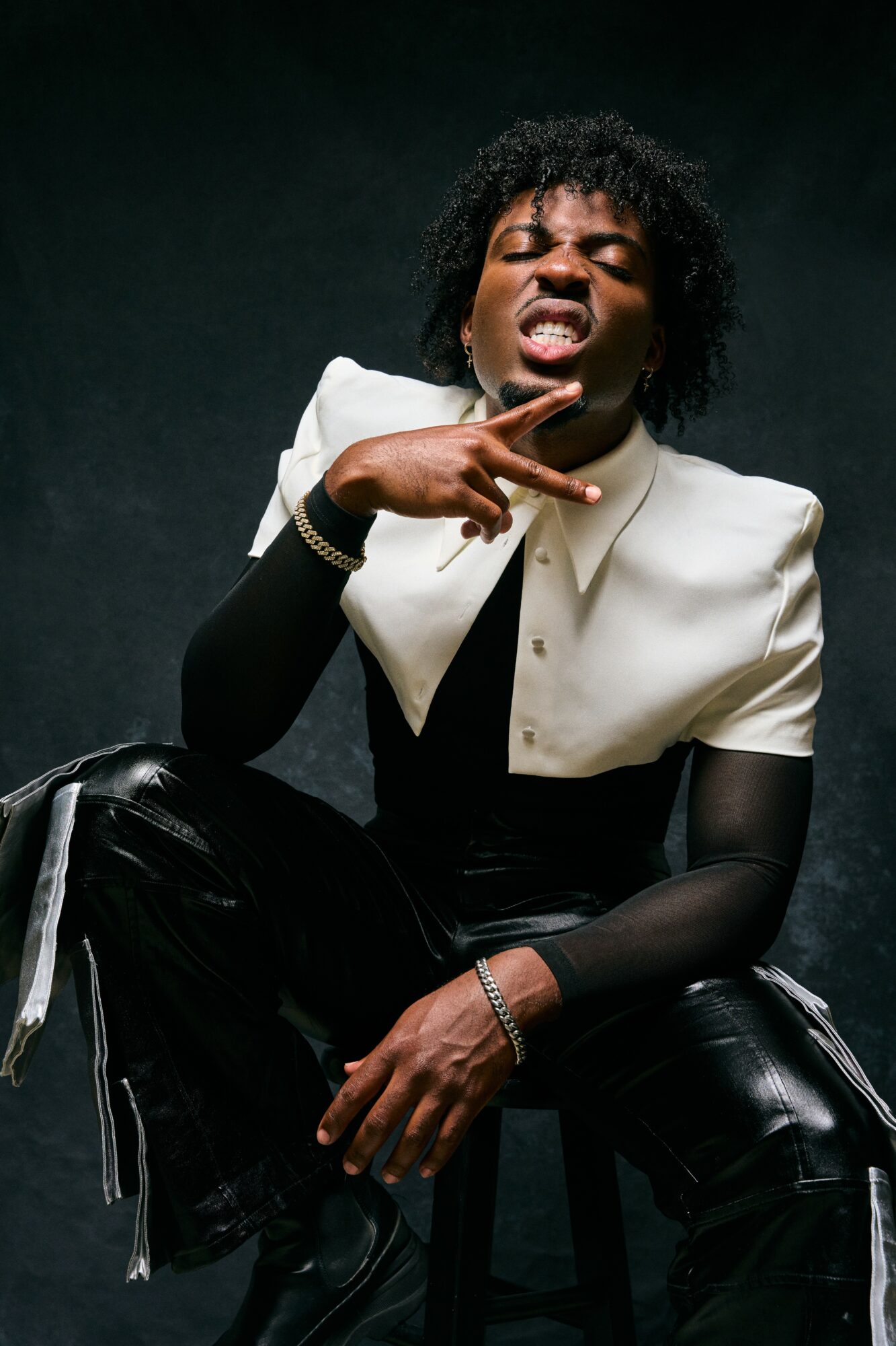 Person with curly hair making a gesture with their hand, wearing a white and black outfit, against a dark background.