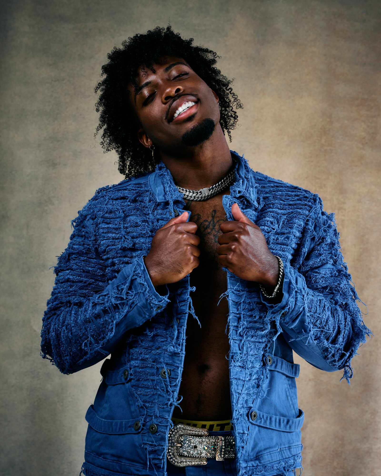Person with curly hair wearing a blue textured jacket, smiling, holding the collar, against a neutral background.