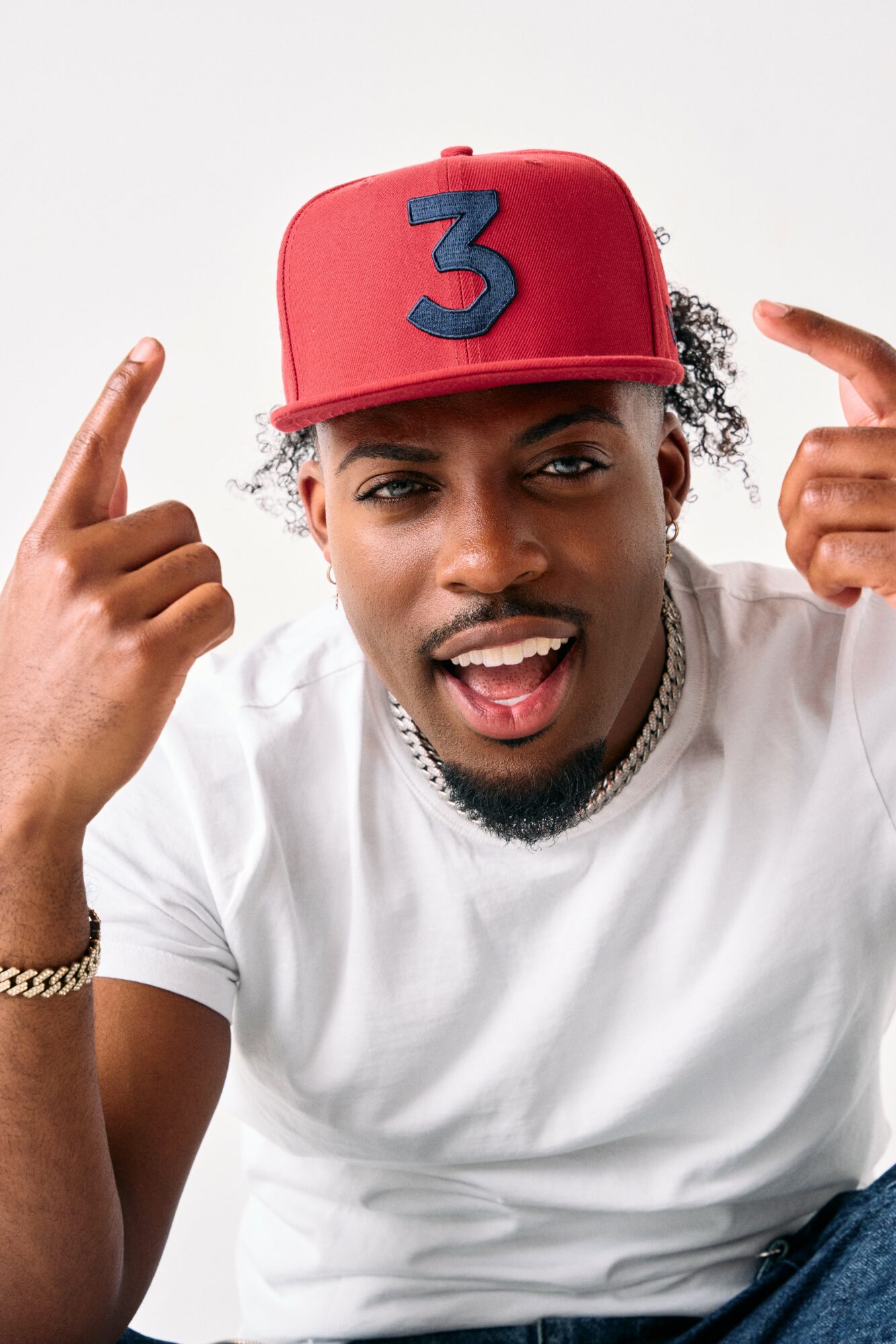 Young man wearing a red cap with a blue number three, pointing at his head, smiling, white background.