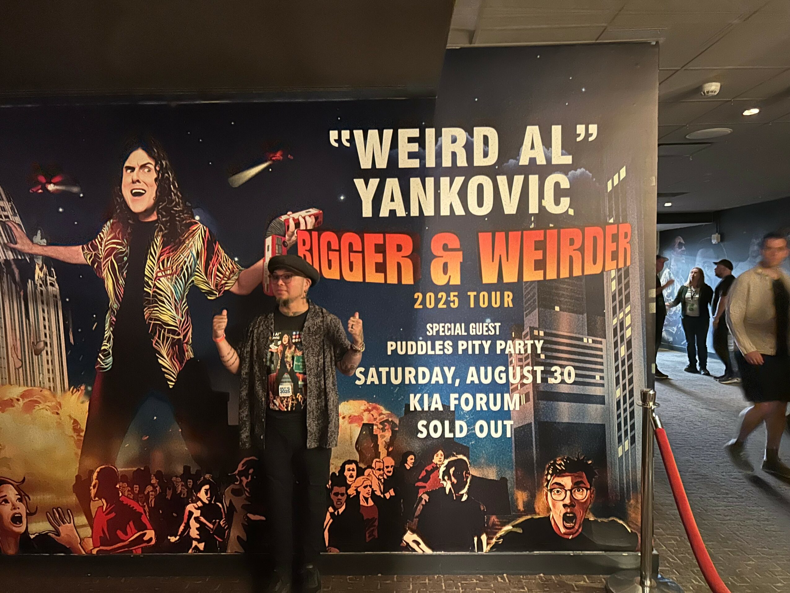 Person standing in front of a large poster for Weird Al Yankovic's 2025 tour, with a woman in a hat and glasses, and a crowd image on the poster.