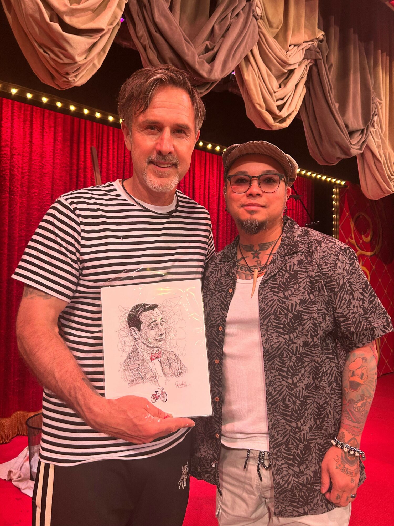 Two men standing on a stage with red curtains, one holding a drawing, the other wearing glasses and a patterned shirt.