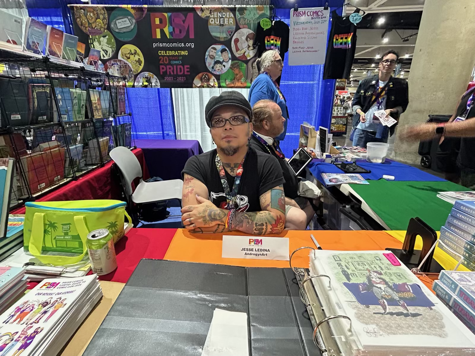 Person with glasses and tattoos sitting at a table with books and a green container, in a colorful event setting.