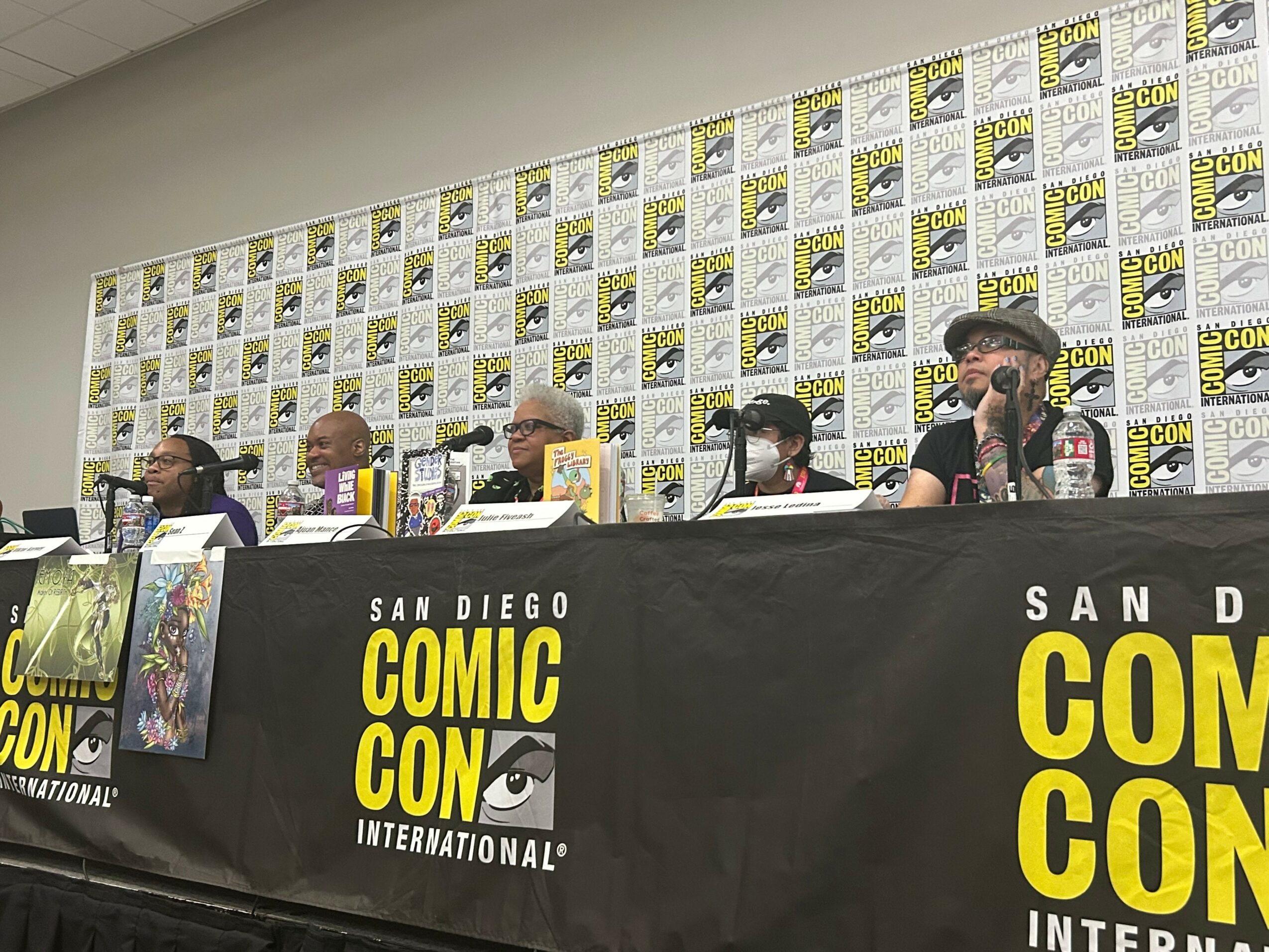 Panel of six people sitting at a table during a San Diego Comic-Con event, with a backdrop of Comic-Con logos.
