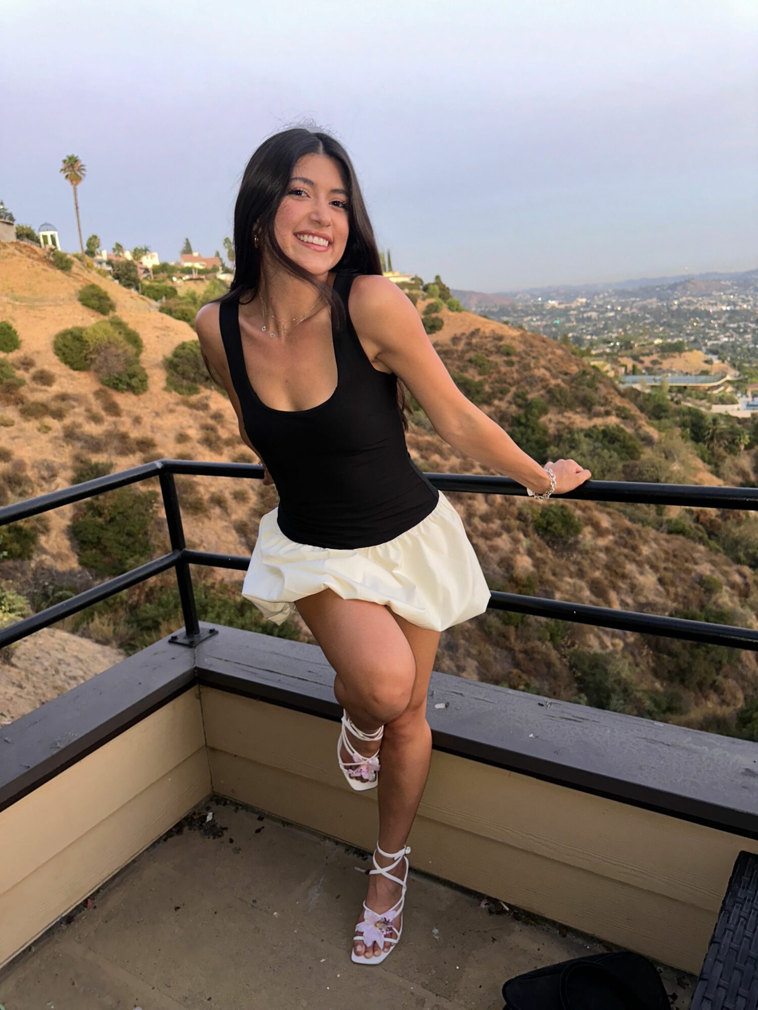 Woman in black top and white skirt standing on balcony with hills and city view behind her.