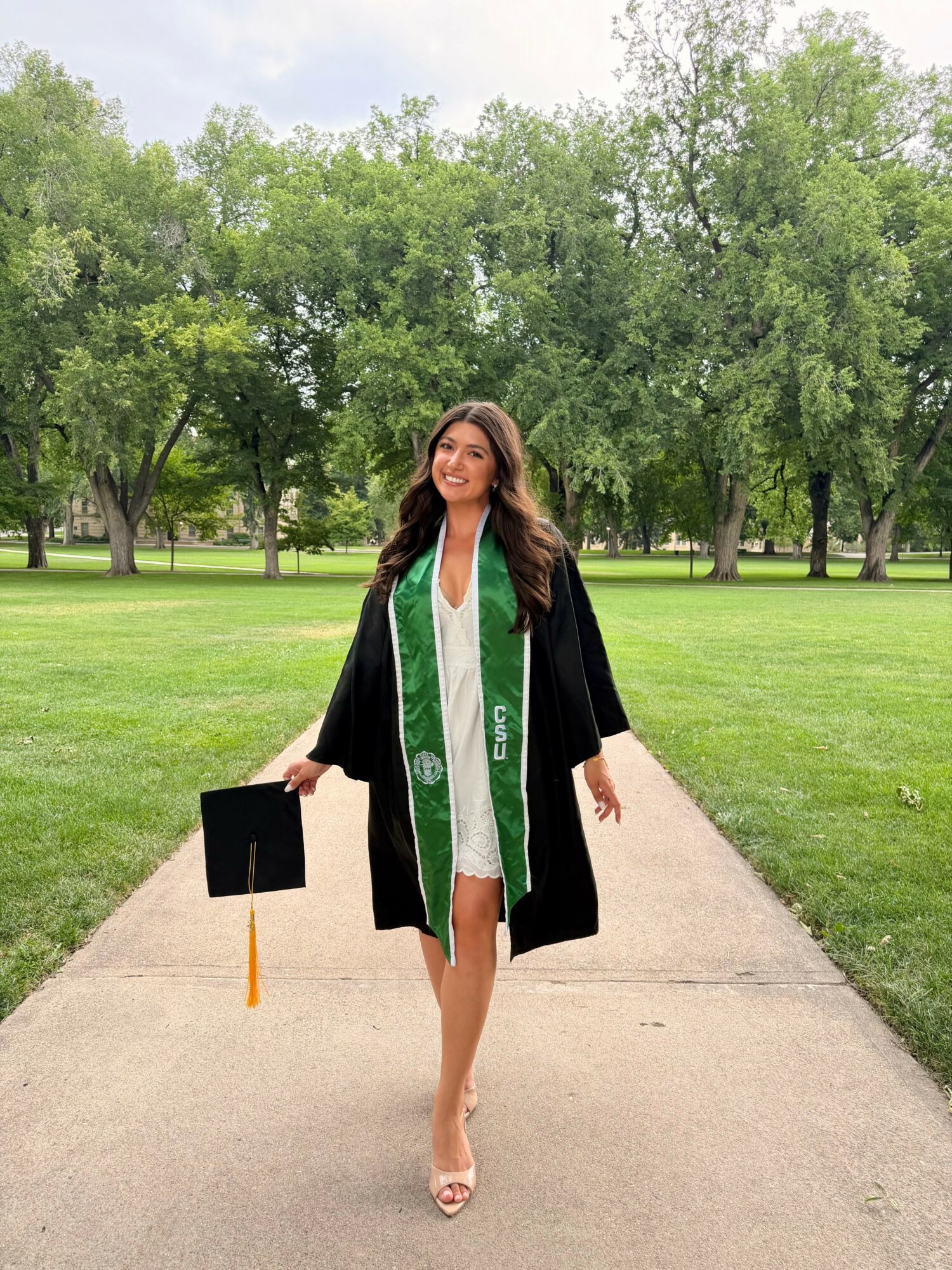 Young woman in graduation gown and cap walking on a park path, smiling, with trees in the background.