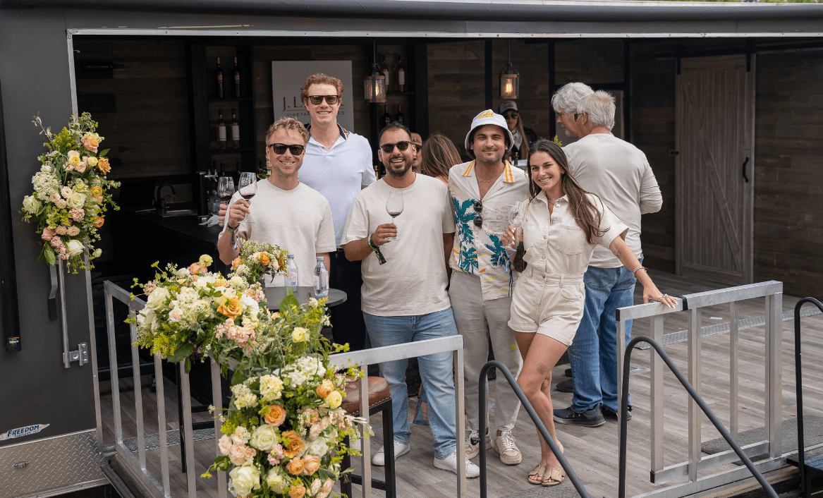 Group of six people standing on a deck with flowers, smiling, holding drinks, in front of a building.