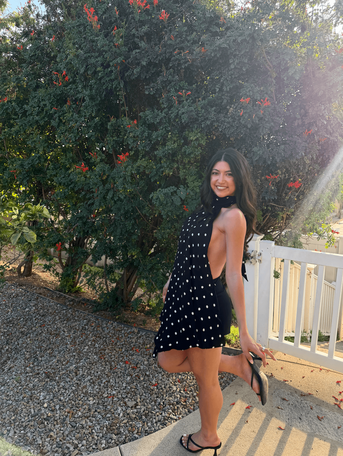 Young woman in a polka dot dress smiling outdoors near a white fence and greenery.