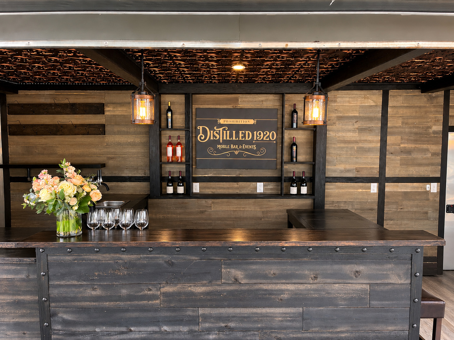 Bar counter with flowers and glasses, wooden wall background, sign reading 'Distilled 1920', warm lighting.