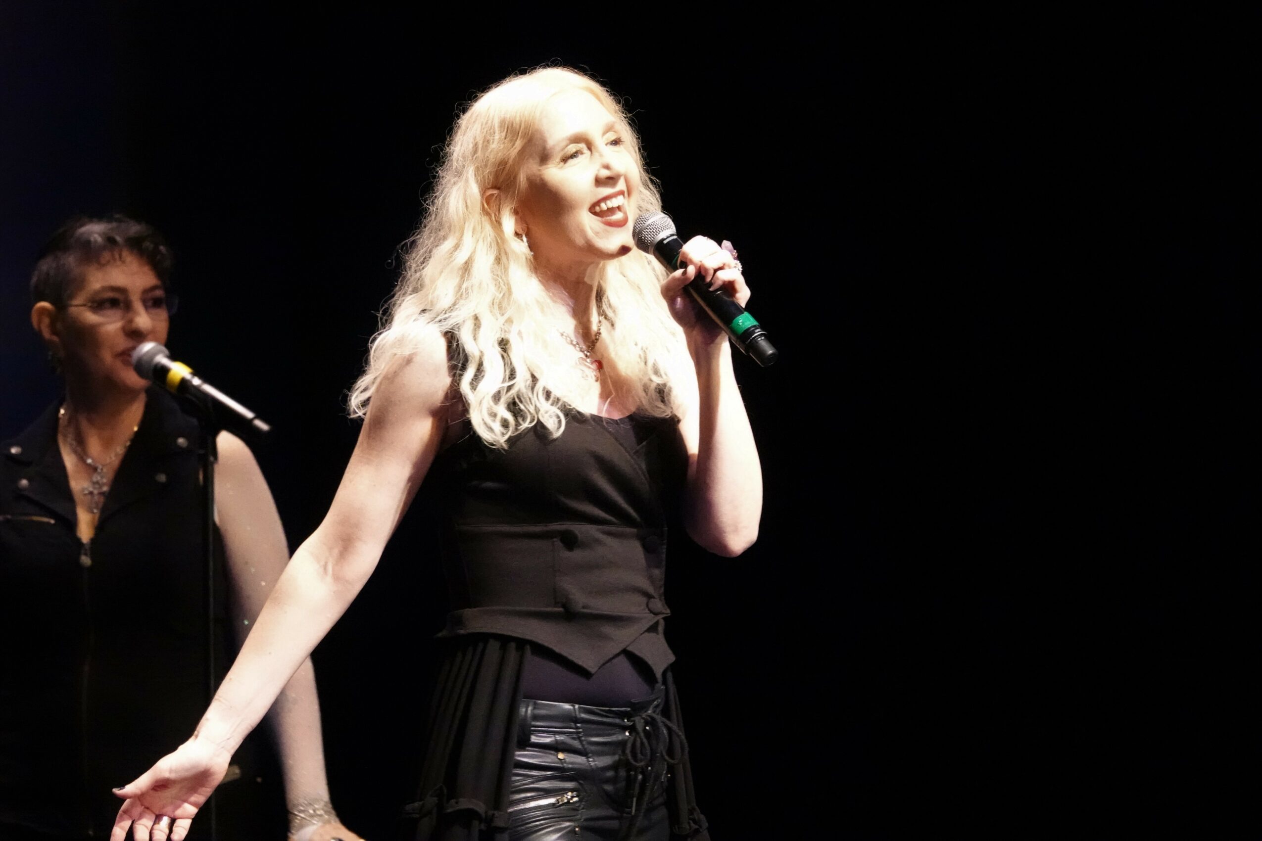 Two women singing on stage, one with long blonde hair holding a microphone, the other with dark hair in the background.