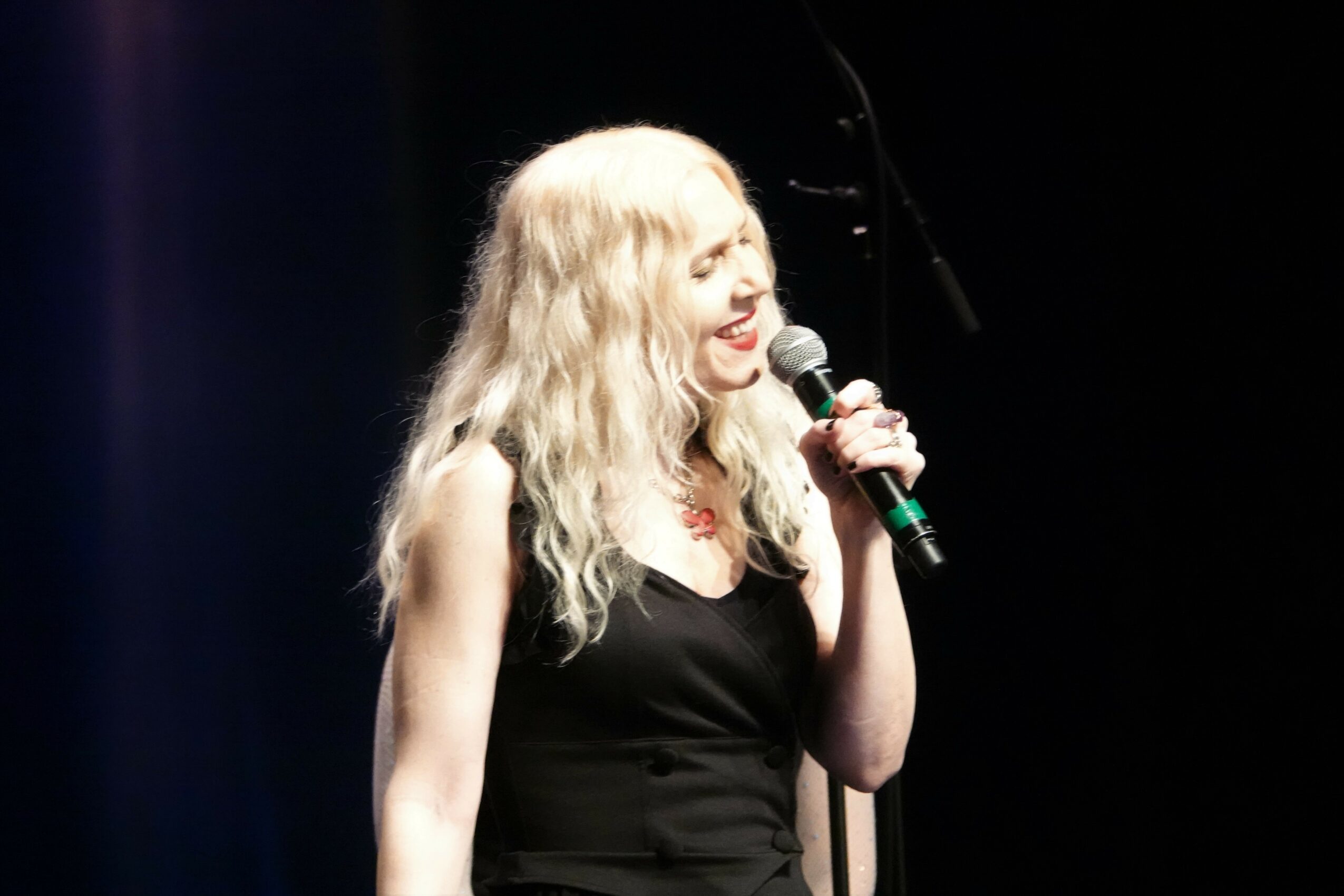 Woman with long blonde hair singing into a microphone on stage, smiling, wearing a black dress.