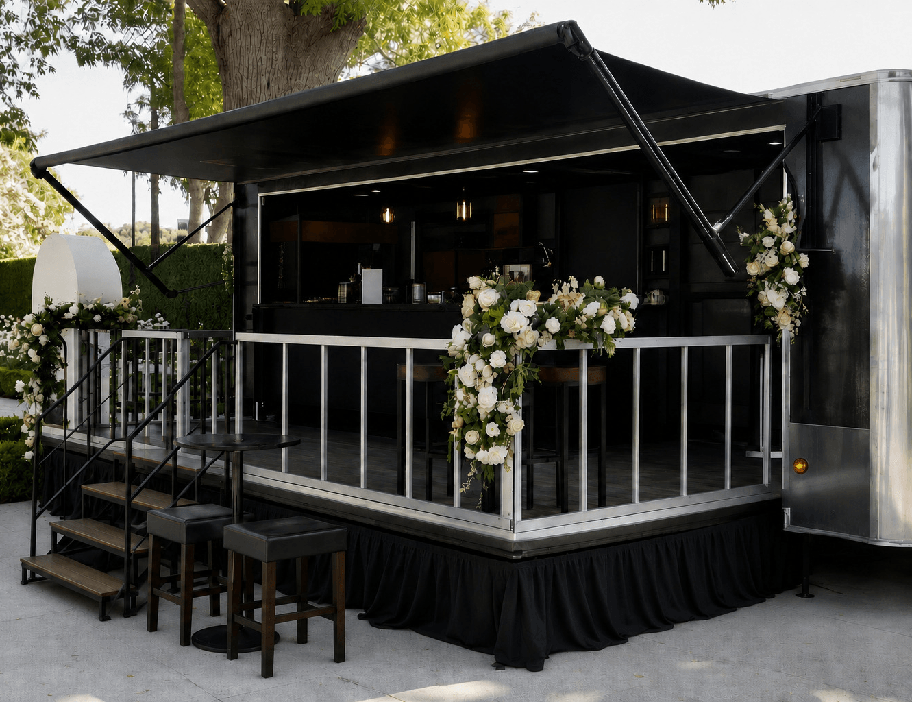 Black mobile bar with open canopy, decorated with white flowers, on a platform with stairs and railing, outdoors with trees.
