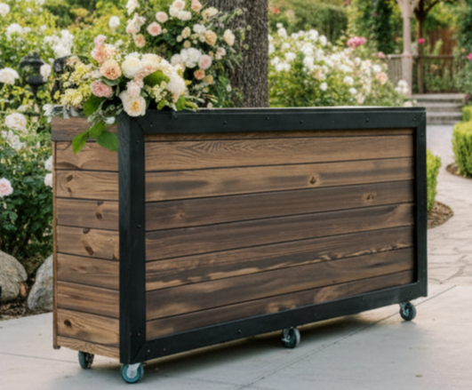 Wooden planter box with flowers on top, outdoors in a garden setting, on wheels, with trees and flowers in background.