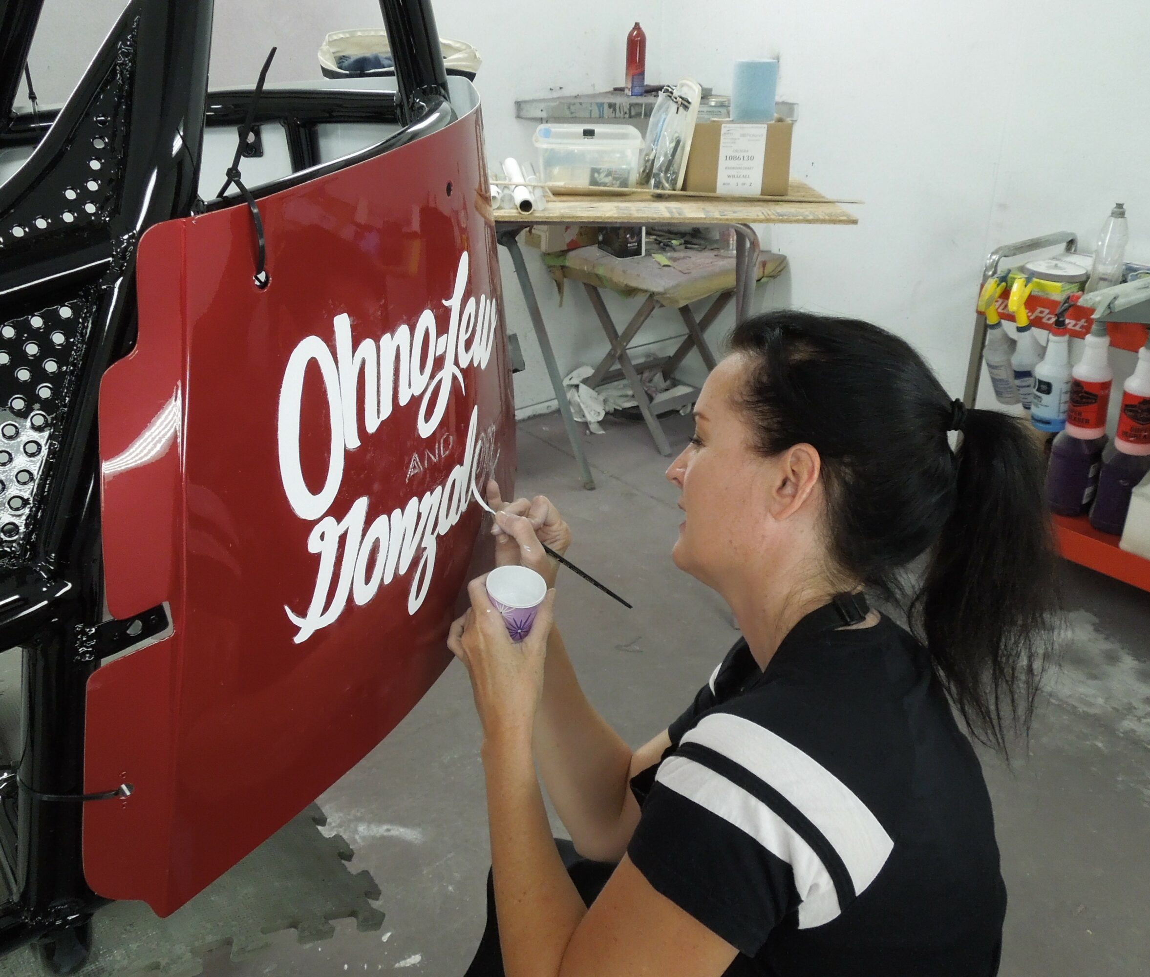 Woman painting white text on a red vehicle in a workshop, holding a small container and brush.