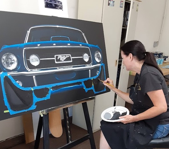 Artist painting a large canvas of a classic car with blue and black colors, sitting on a stool in a room.