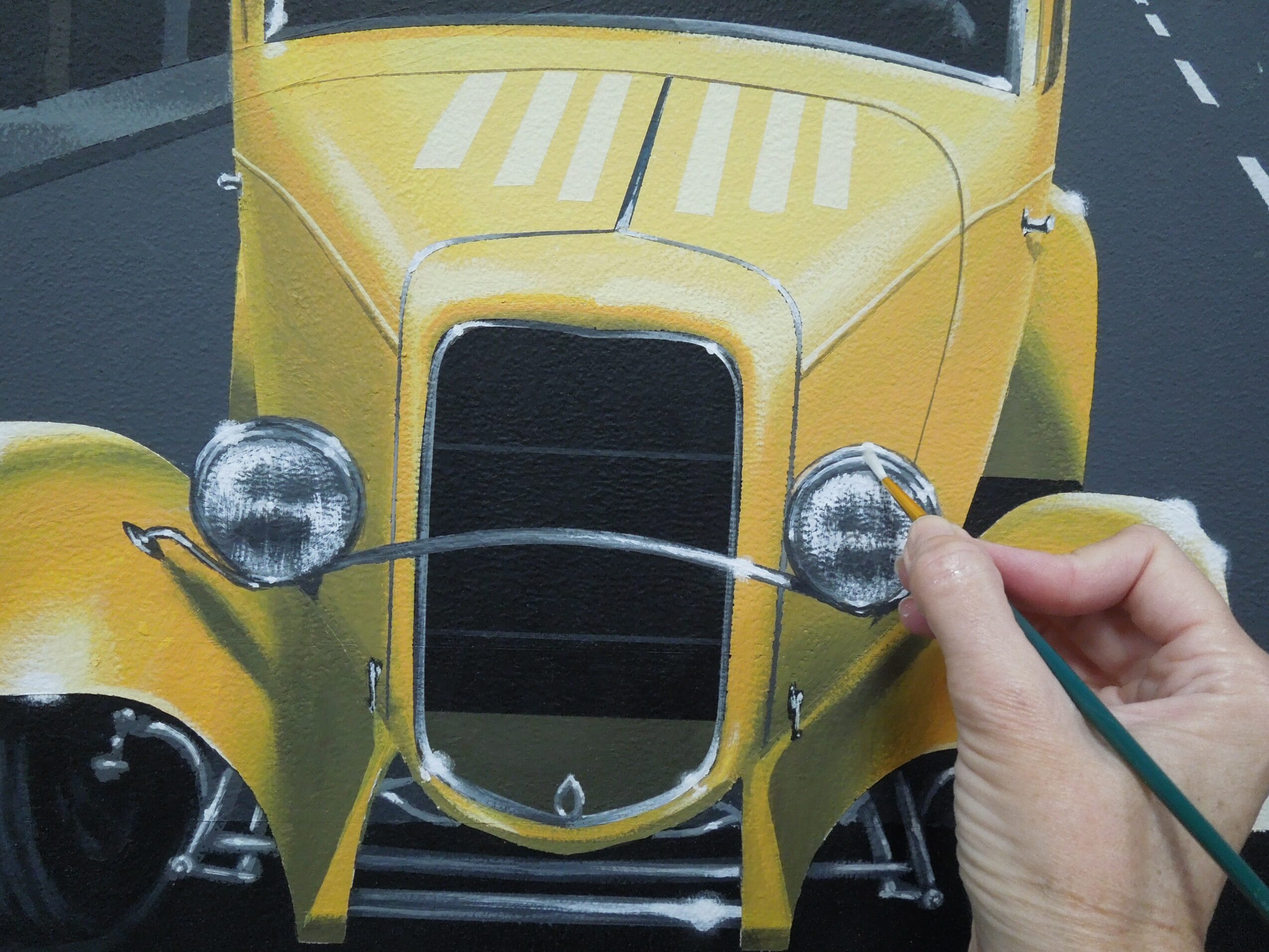 Person holding a paintbrush near the front of a yellow vintage car with round headlights.