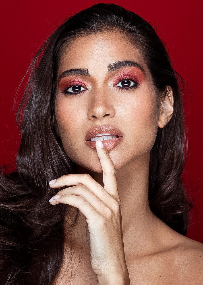 Close-up of a woman with dark hair, red eyeshadow, and lips, holding her finger to her lips against a red background.