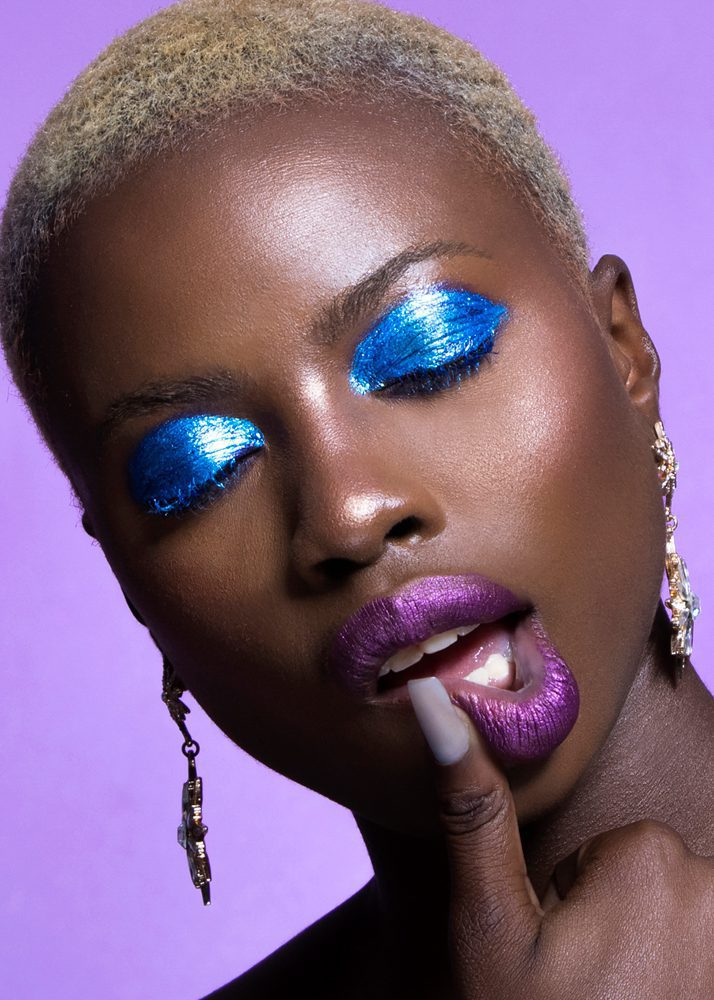 Close-up of a person with short blonde hair, vibrant blue eyeshadow, purple lipstick, and earrings, against a purple background.