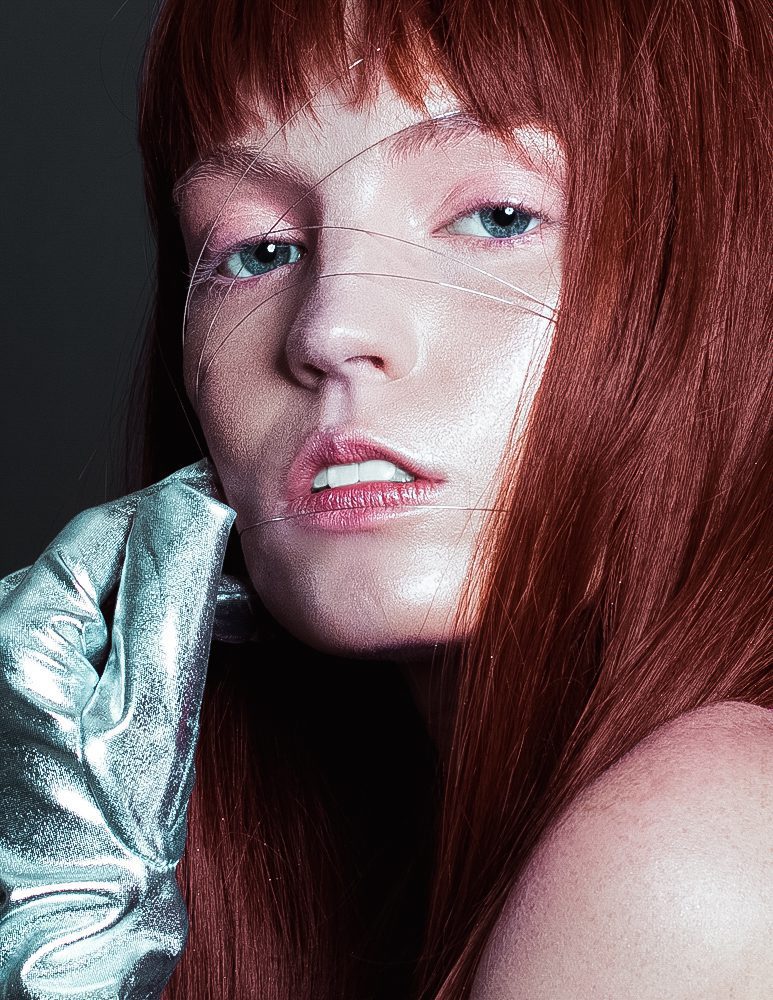 Close-up of a woman with red hair, blue eyes, and light makeup, wearing a shiny silver glove, looking at the camera.