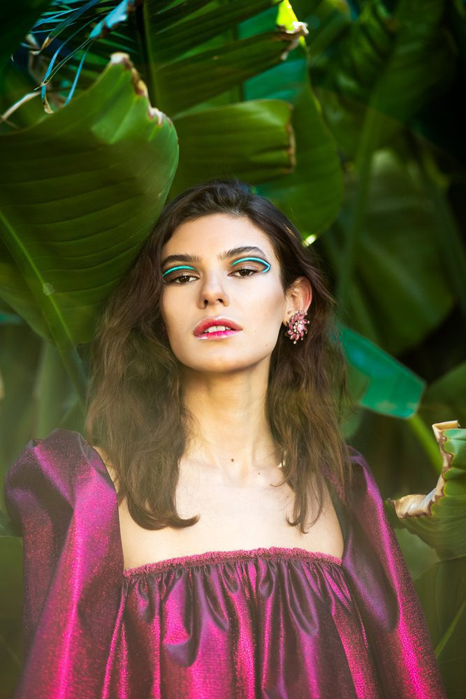 Woman with makeup and earrings in front of large green leaves, wearing a pink off-shoulder top.