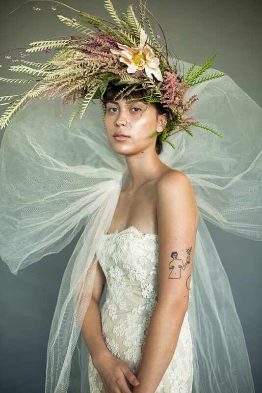 Woman with a large floral headpiece and sheer fabric, wearing a strapless dress, standing against a plain background.