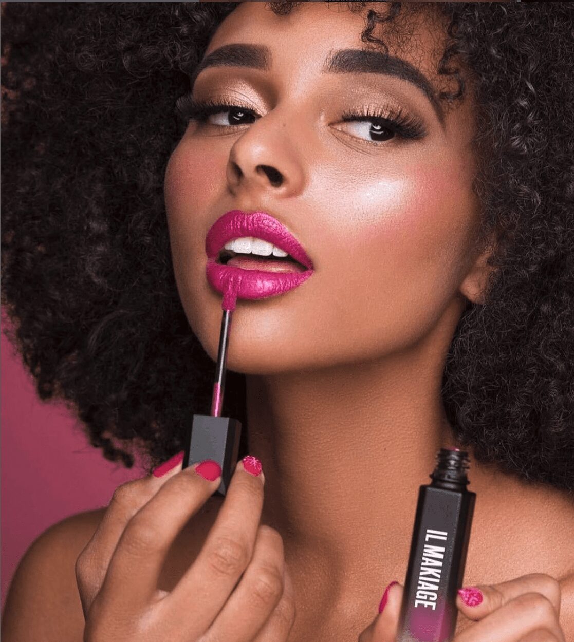 Woman applying pink lipstick with a brush, holding a lipstick tube, with curly hair and makeup, pink background.
