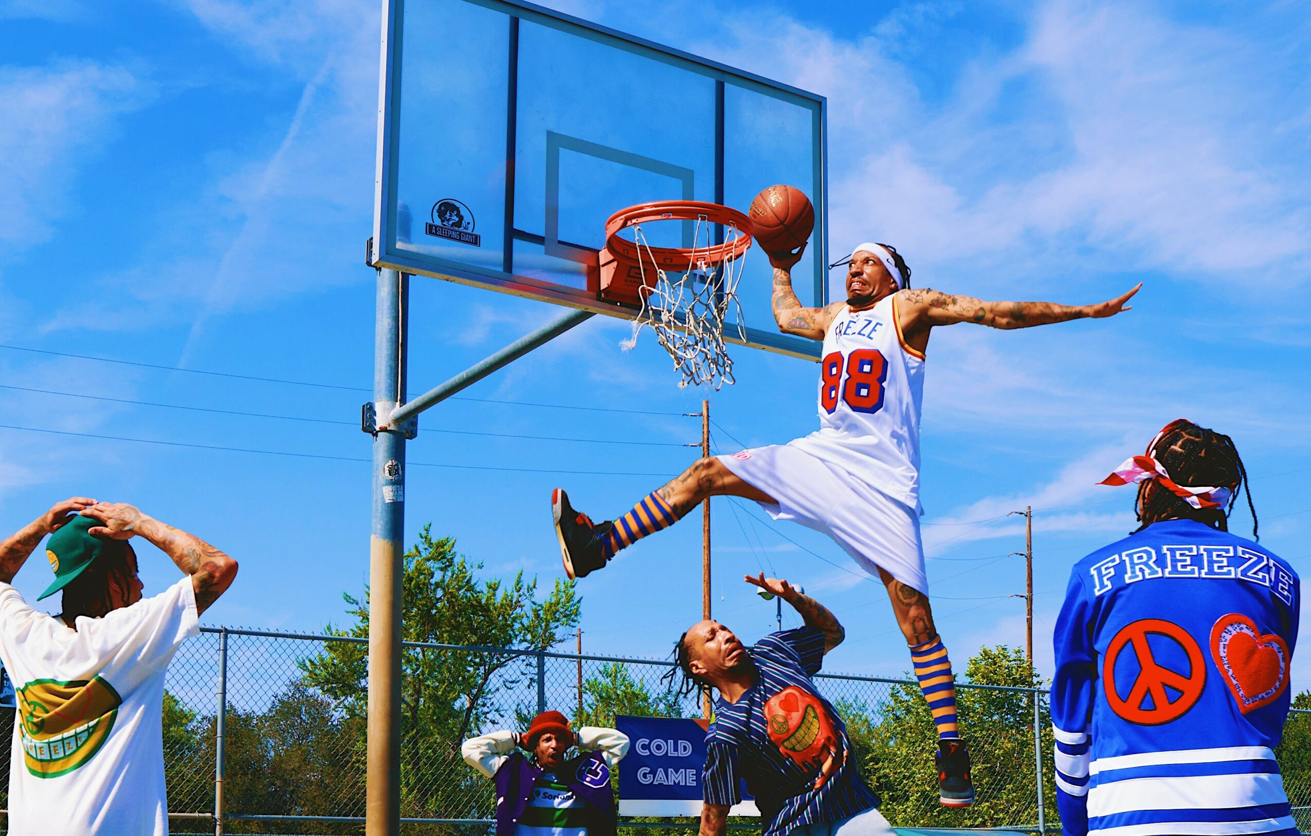Person in a white jersey jumps to dunk a basketball, others watch outdoors on a sunny day.