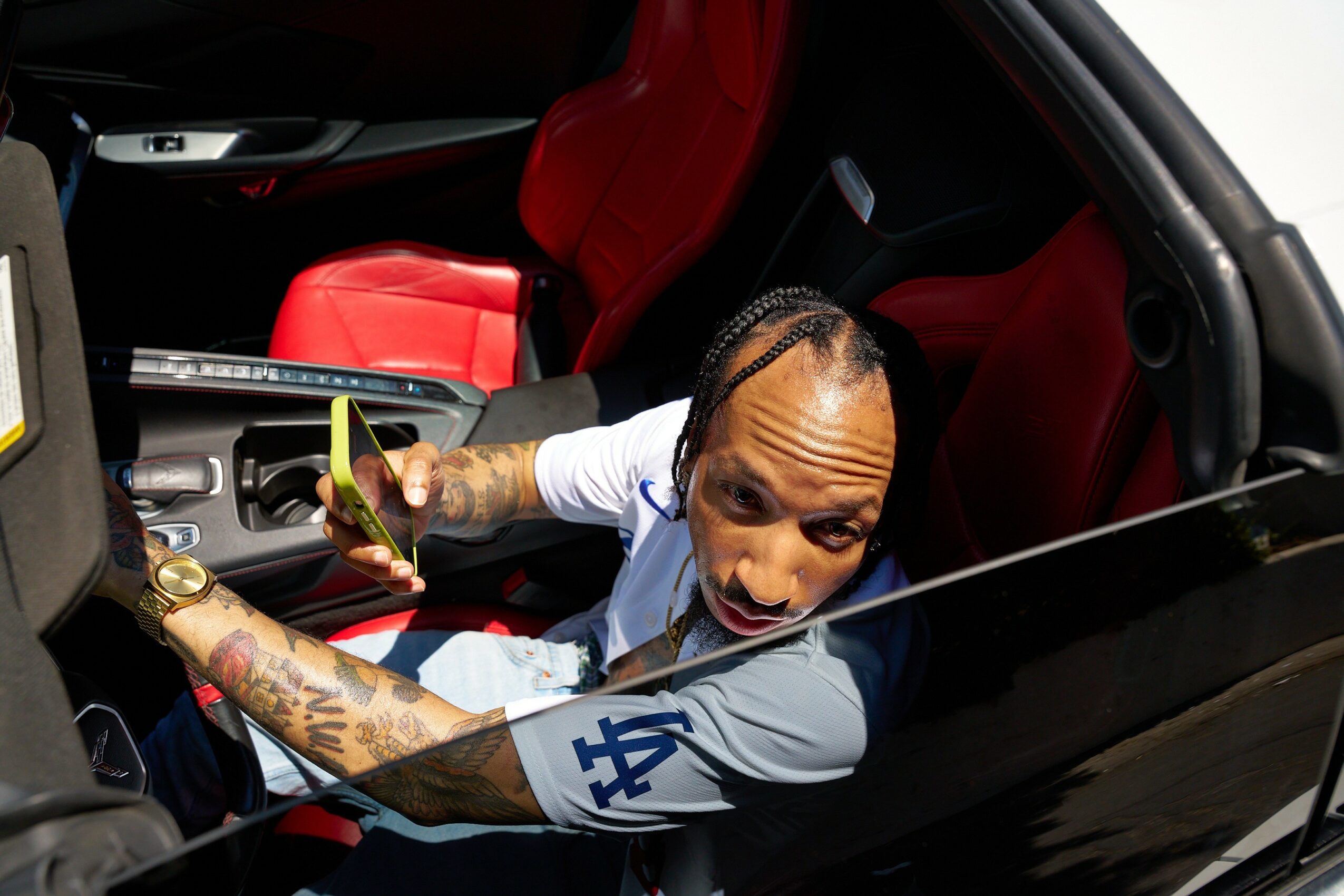 Man with tattoos in a white and gray Los Angeles Dodgers jersey sits in a car with red interior, looking up at the camera.