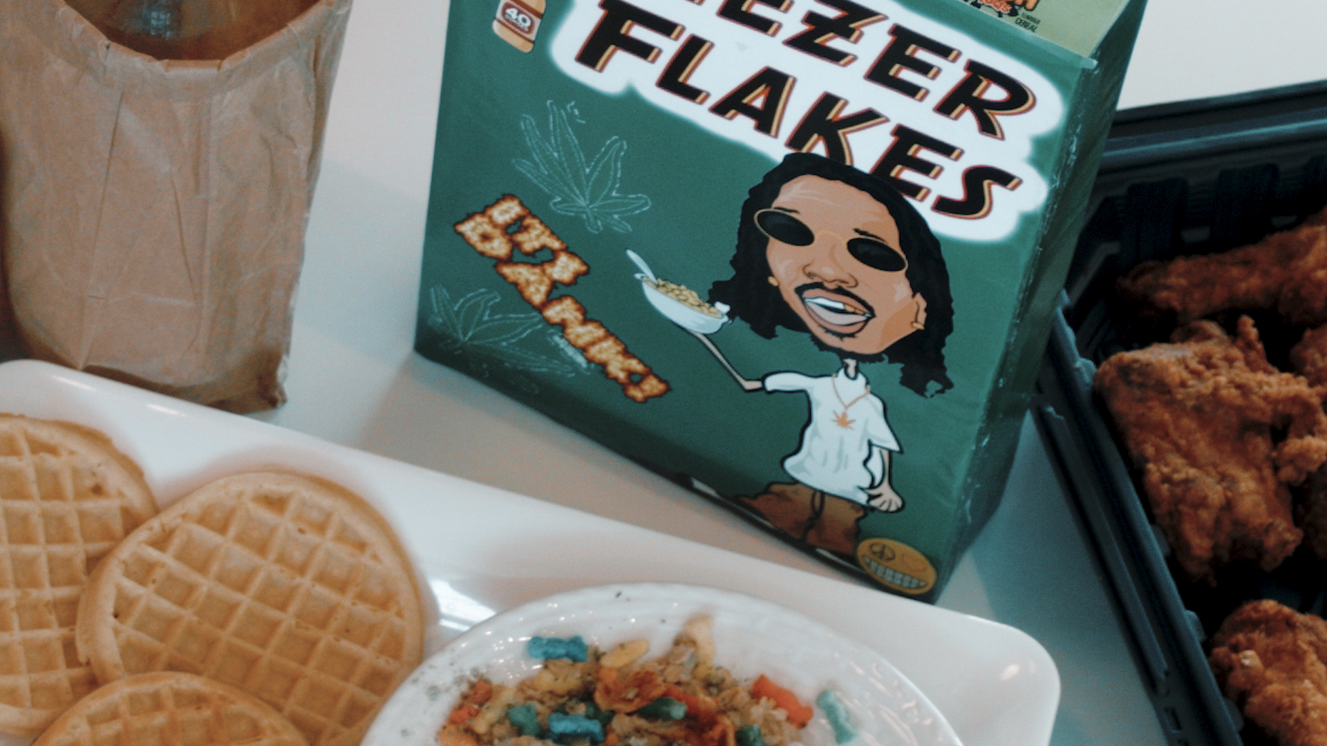 Box of cereal, waffles, and a bowl of cereal on a table, with fried chicken in the background.