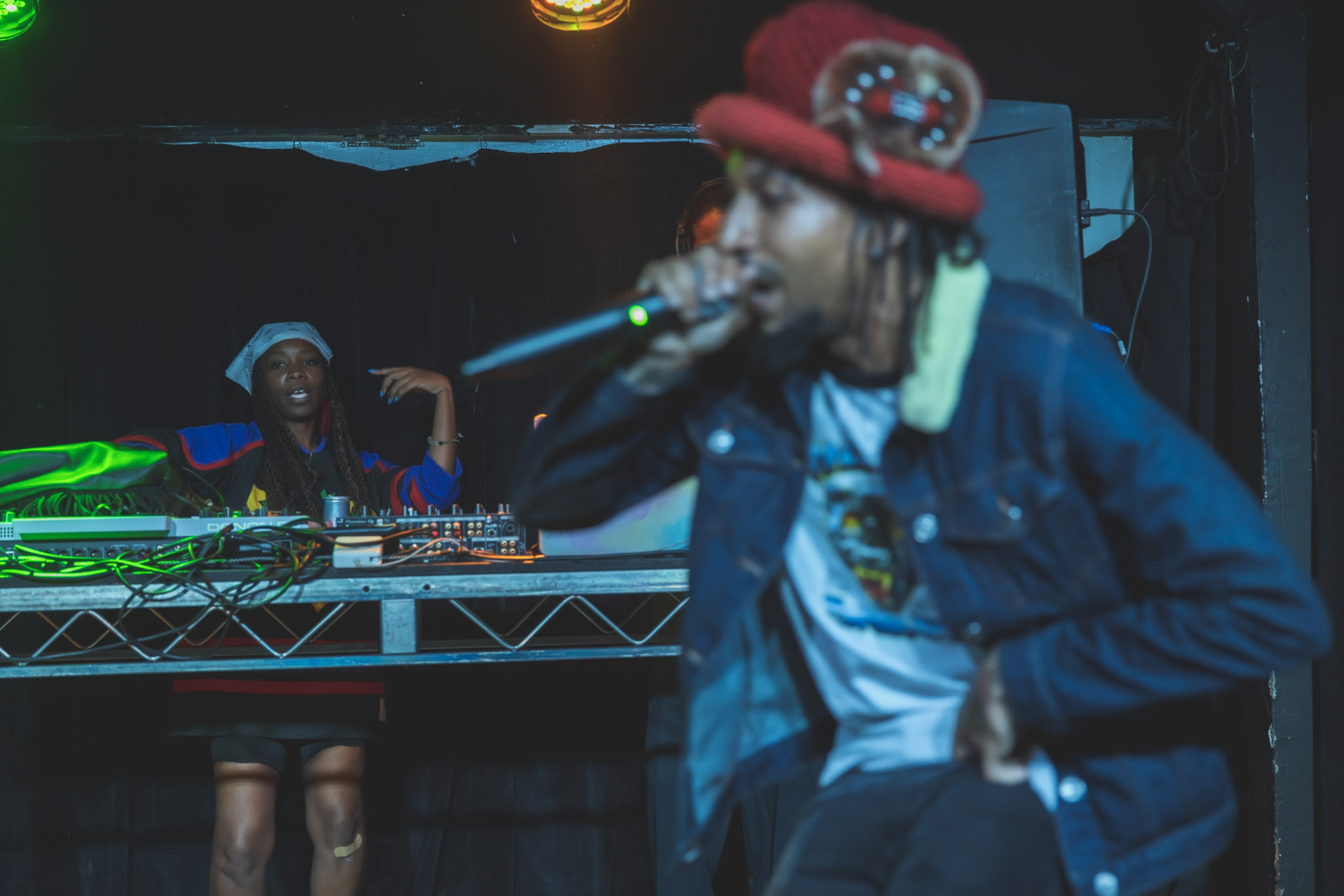 Person wearing a red hat and denim jacket singing into a microphone on stage, with a DJ in the background.