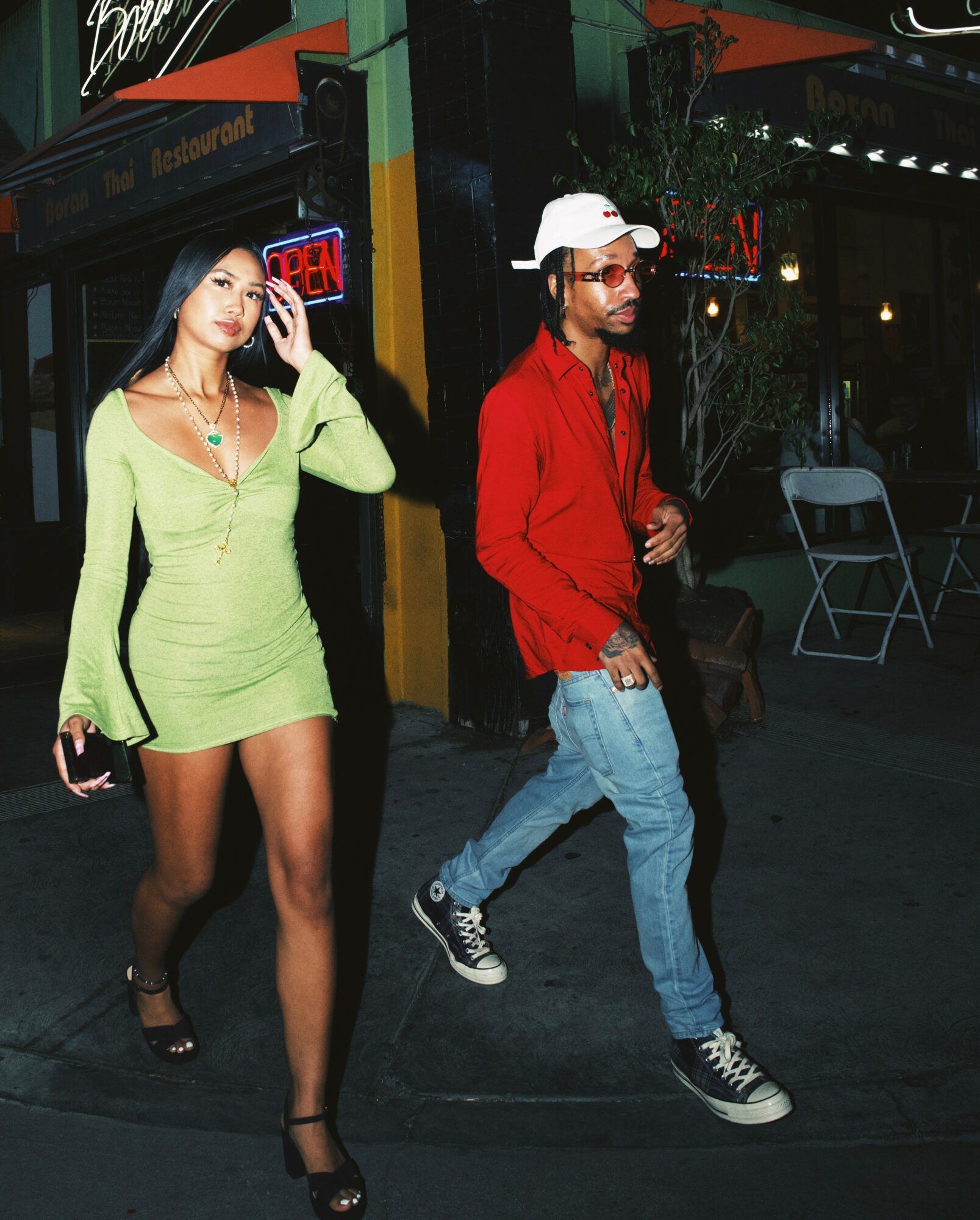 A woman in a lime green dress and a man in a red jacket walk on a city street at night.